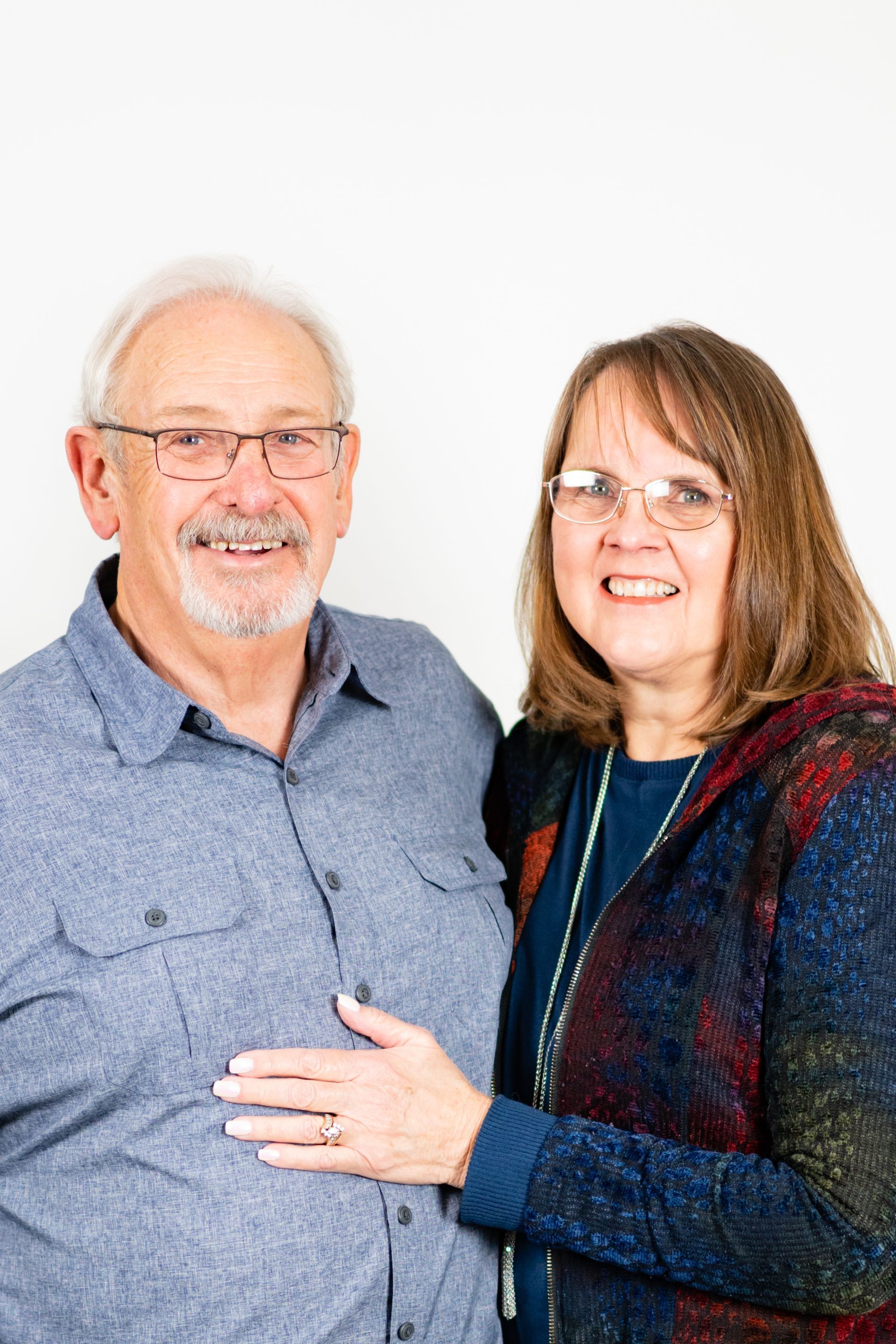 Older couple, man in blue shirt, woman in patterned coat, both smiling, standing together.