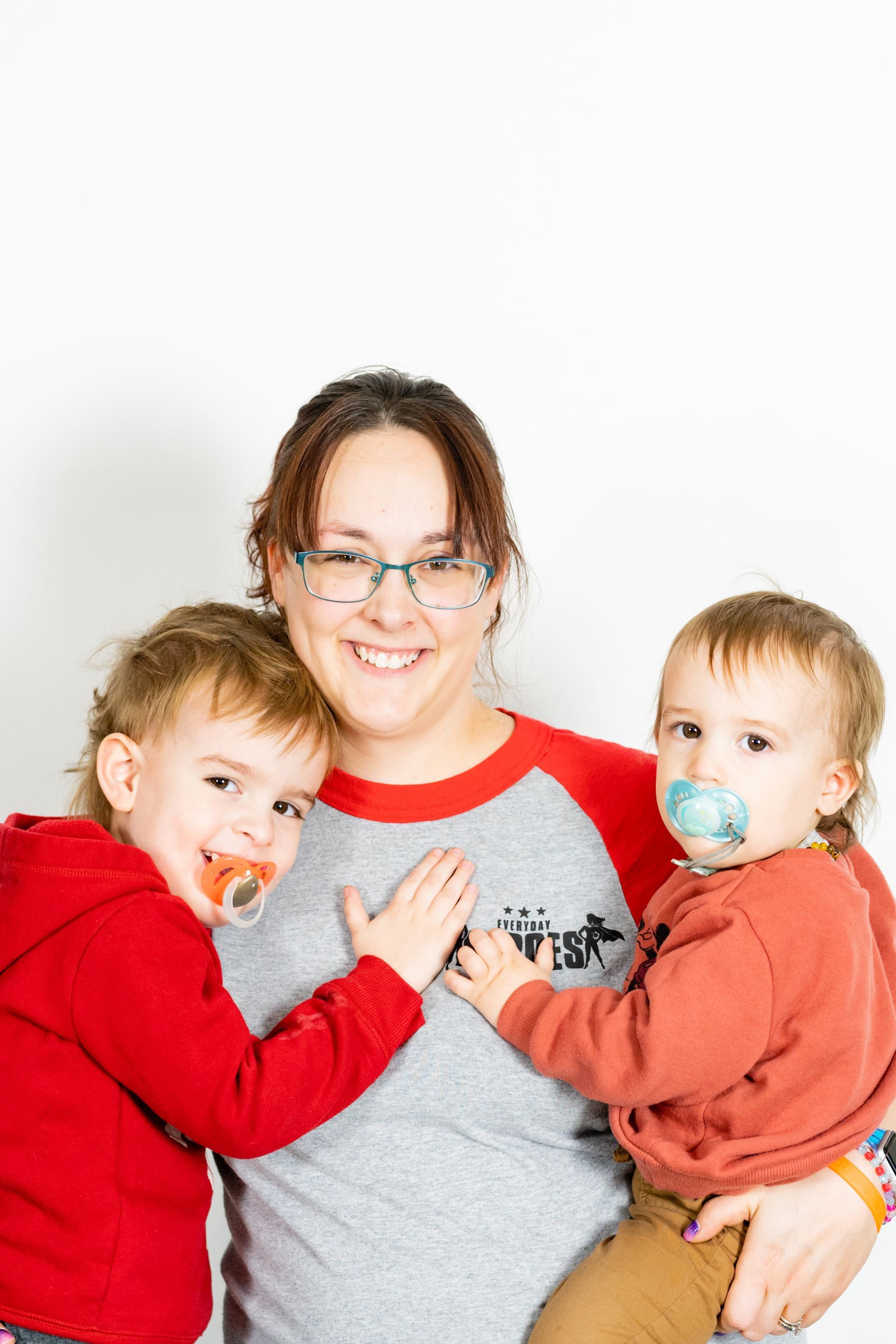Woman in glasses smiles holding two toddlers with pacifiers, standing in front of a white background.