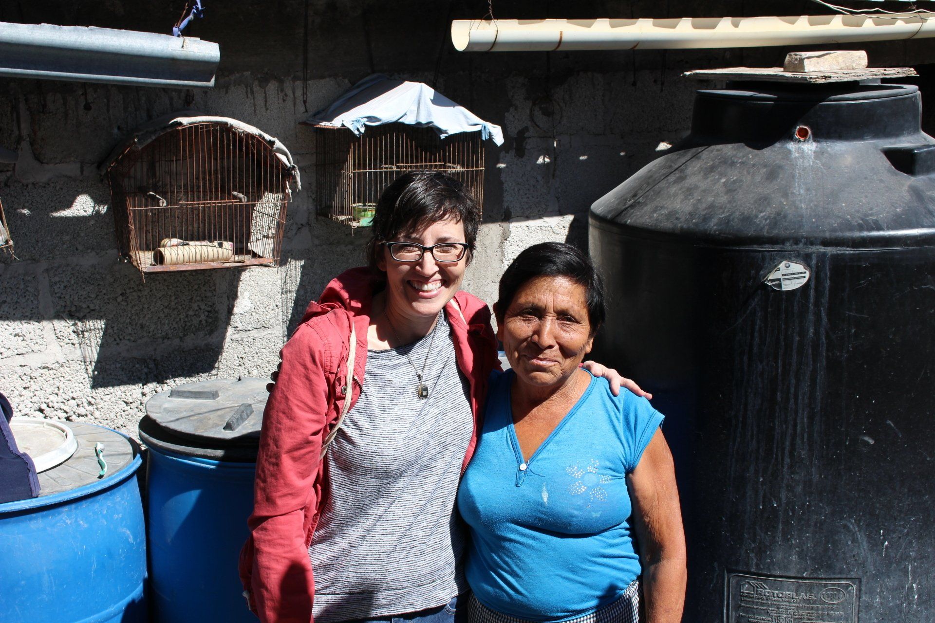 Woman in red jacket and woman in blue shirt pose together by wall with bird cages and large water tank.