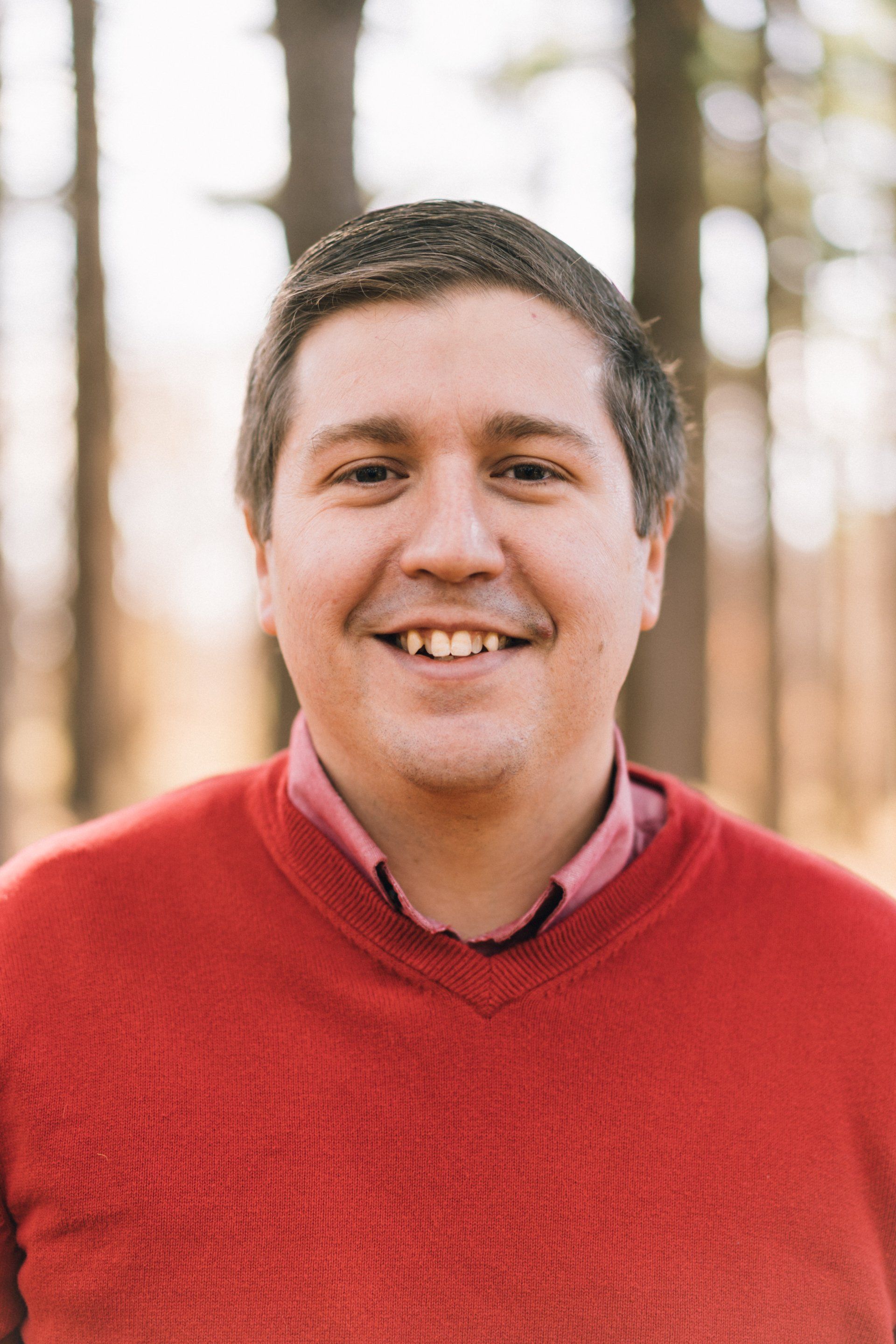 Man smiling, wearing red sweater and pink collared shirt, in a wooded area.