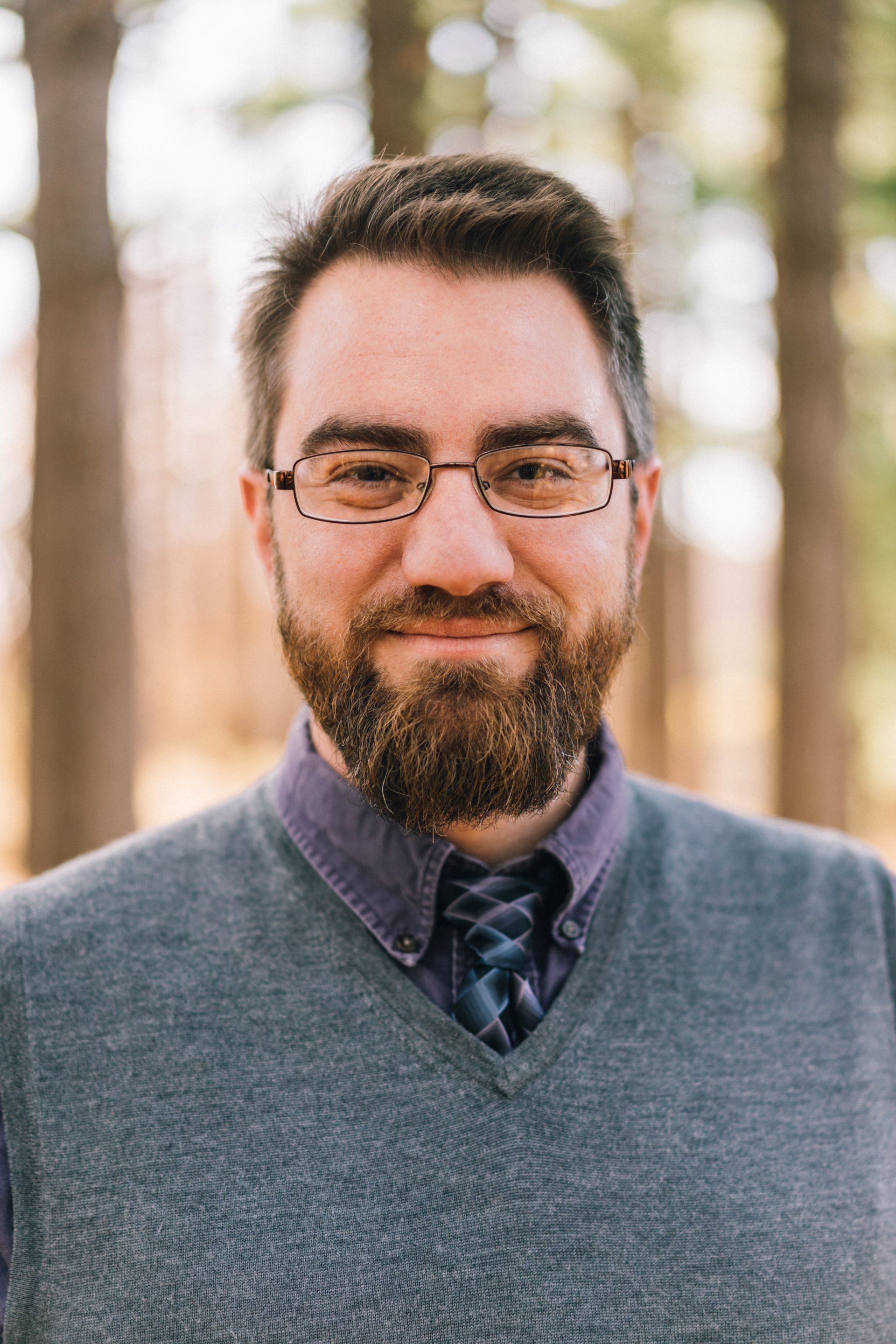 Man with beard and glasses wearing a blue sweater, outdoors with trees in the background.