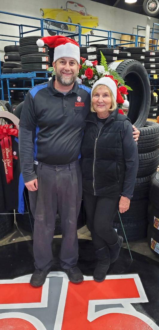 Two people wearing Santa hats stand side-by-side in a tire shop, smiling in front of stacks of tires | 55 Tires and Service
