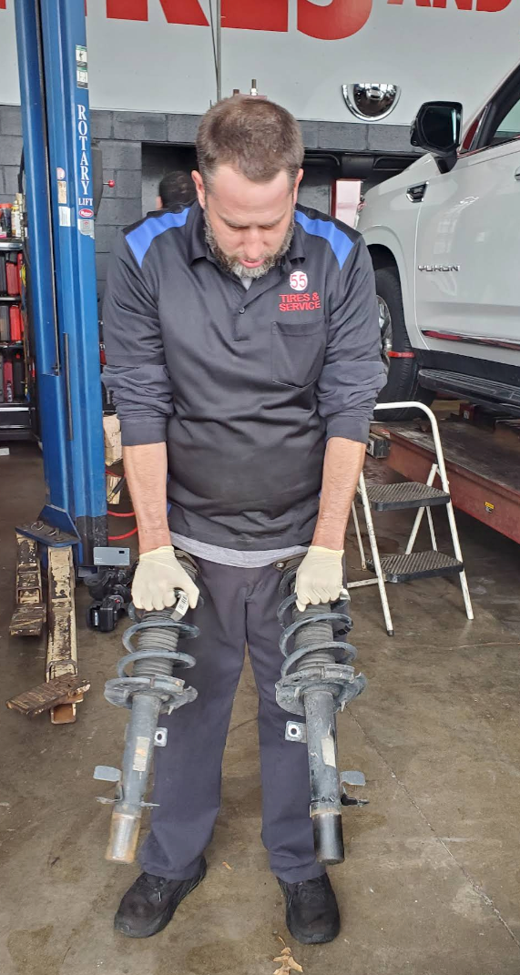 A mechanic in a garage workshop holds two removed vehicle shock absorbers in his gloved hands