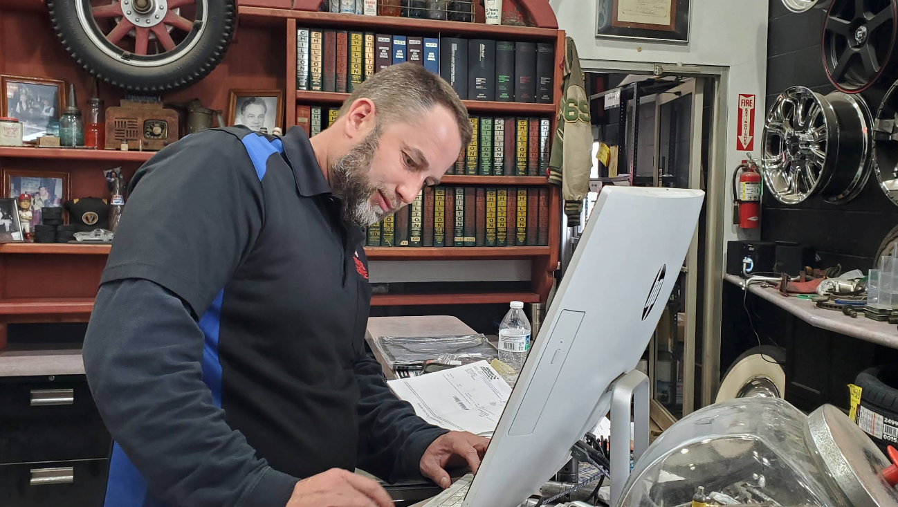 A person with a beard wearing a dark blue work shirt works on a computer in an auto repair shop with shelves of books | 55 Tires and Service