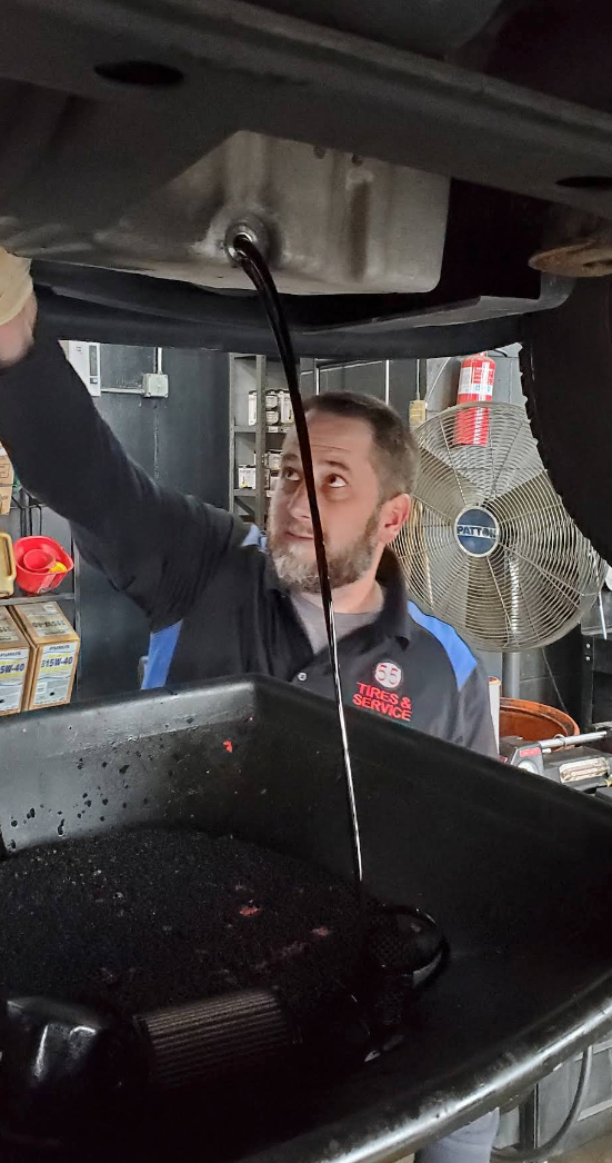 A mechanic works under a lifted vehicle, draining dark, used oil into a collection pan in an automotive shop | 55 Tires and Service