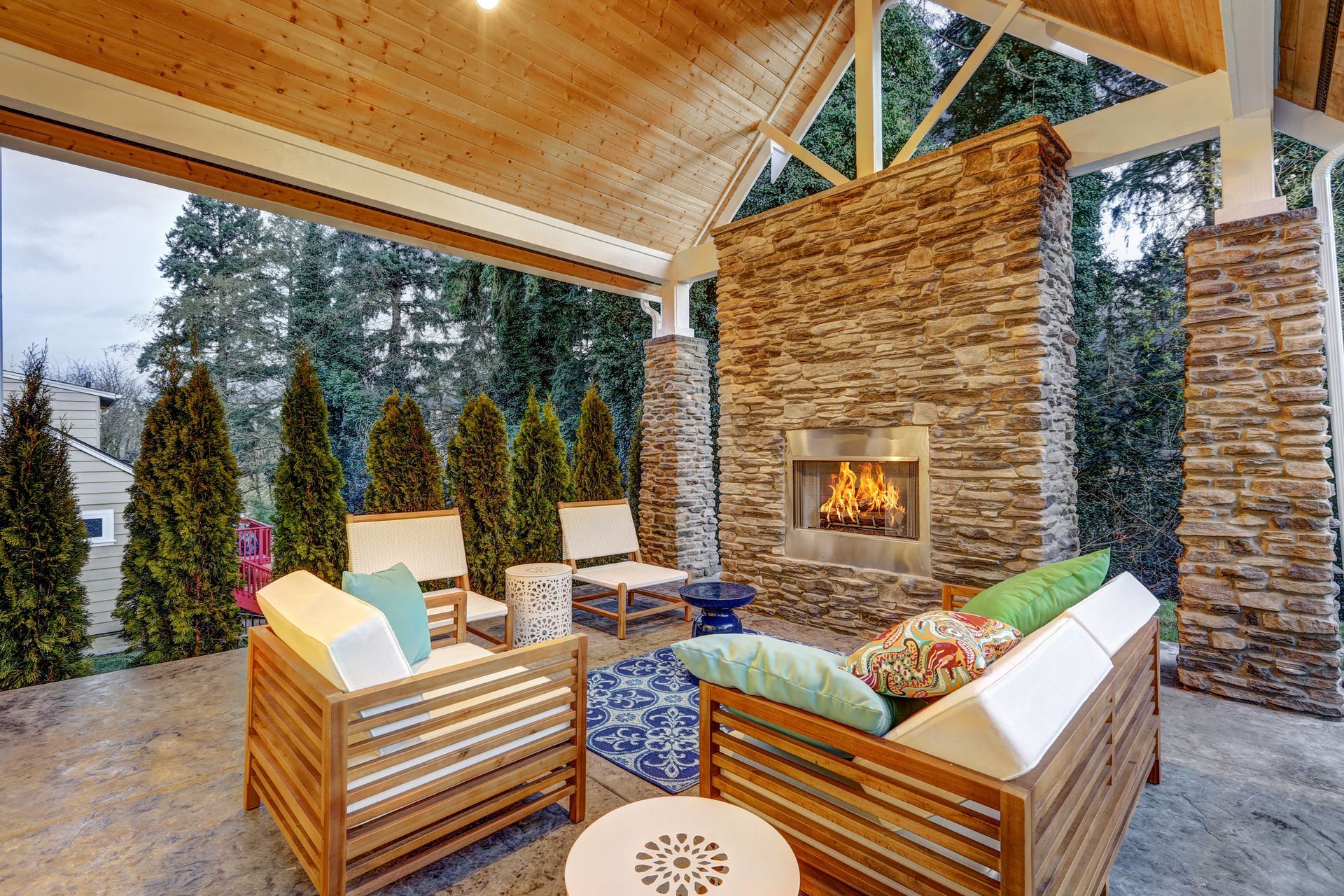 Covered patio with fireplace, seating area, wooden beams, stone fireplace, and surrounding trees.