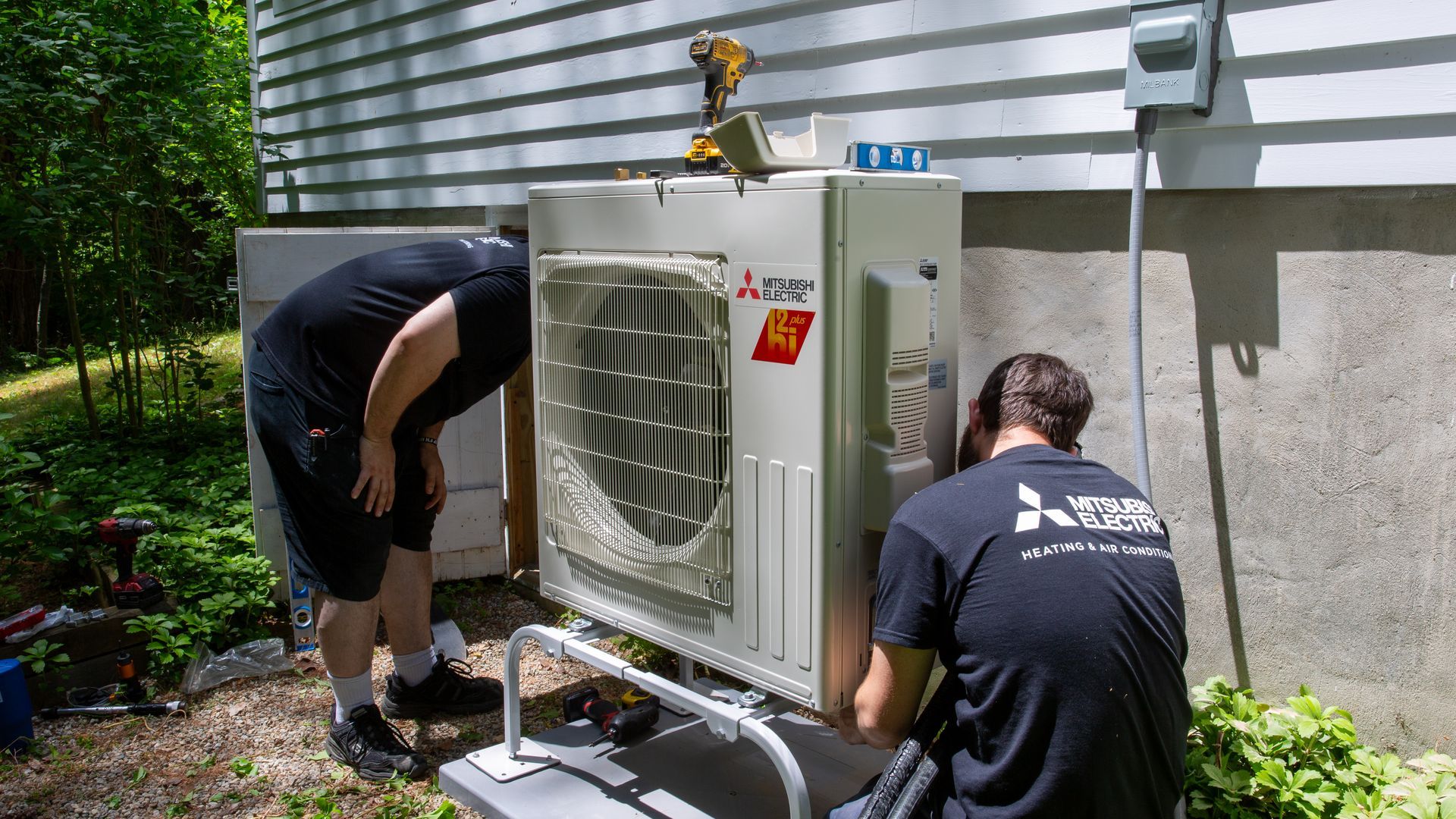 Two Pfluke Home Services technician installing a Mitsubishi heat pump unit on a building exterior.