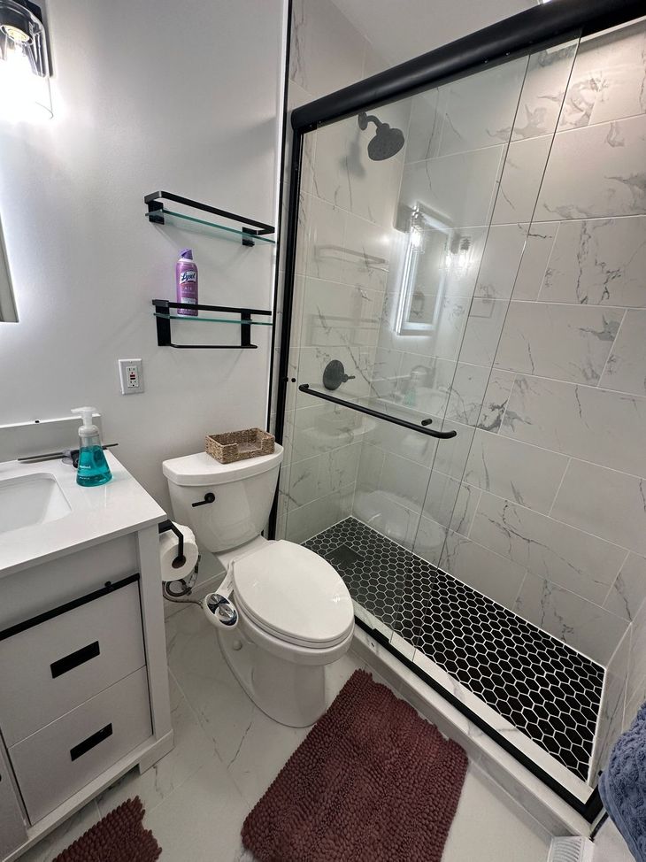 Small bathroom with toilet, sink, glass shower, and burgundy rug.