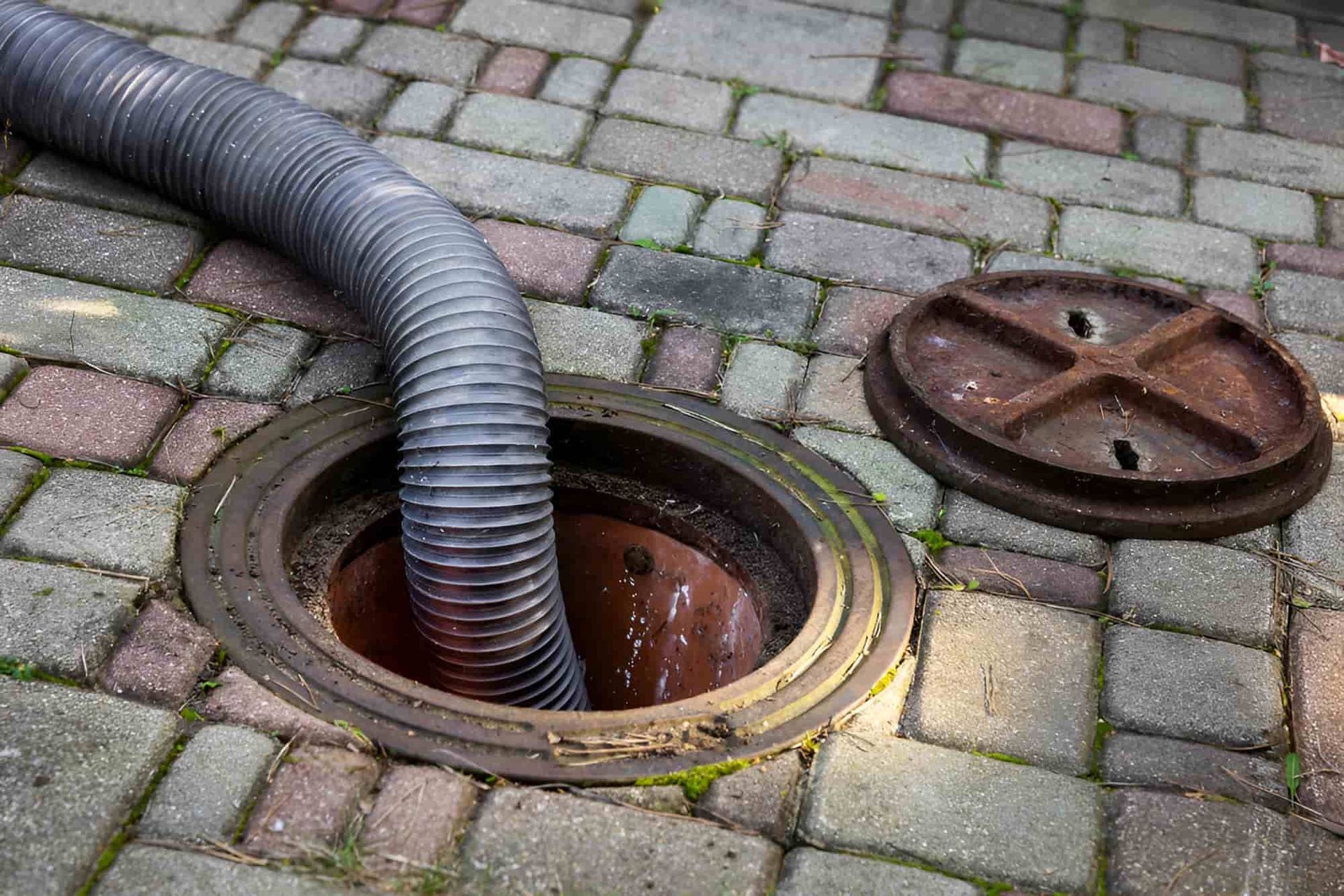 A Hose is Coming Out of a Manhole Cover on a Brick Sidewalk — Simpson's Plumbing & Maintenance Service in Dakabin, QLD