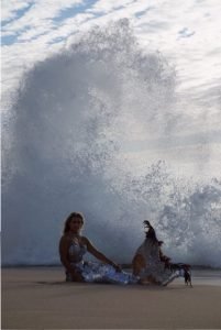 A woman is sitting on the beach in front of a wave.