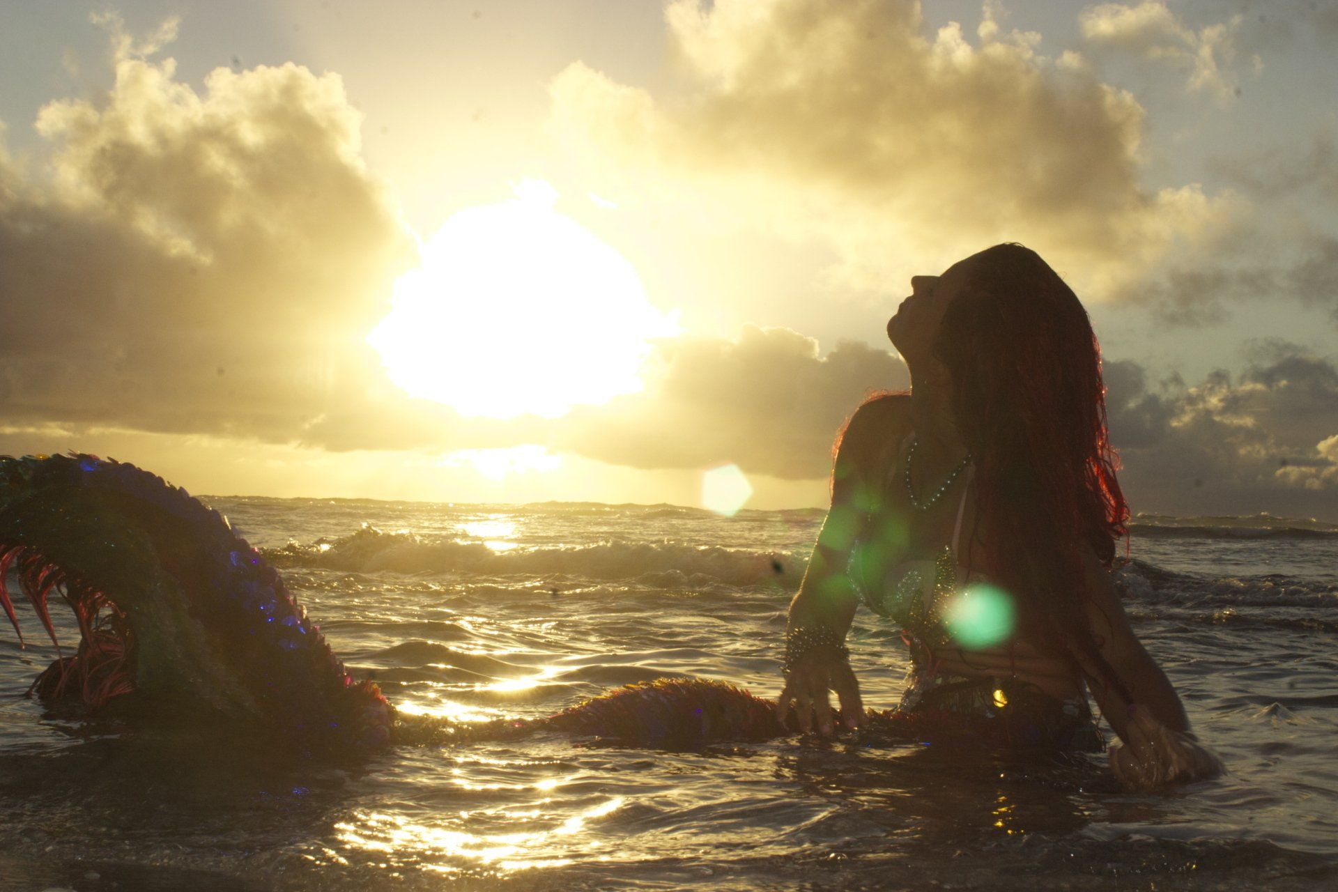 A mermaid is sitting in the ocean at sunset.
