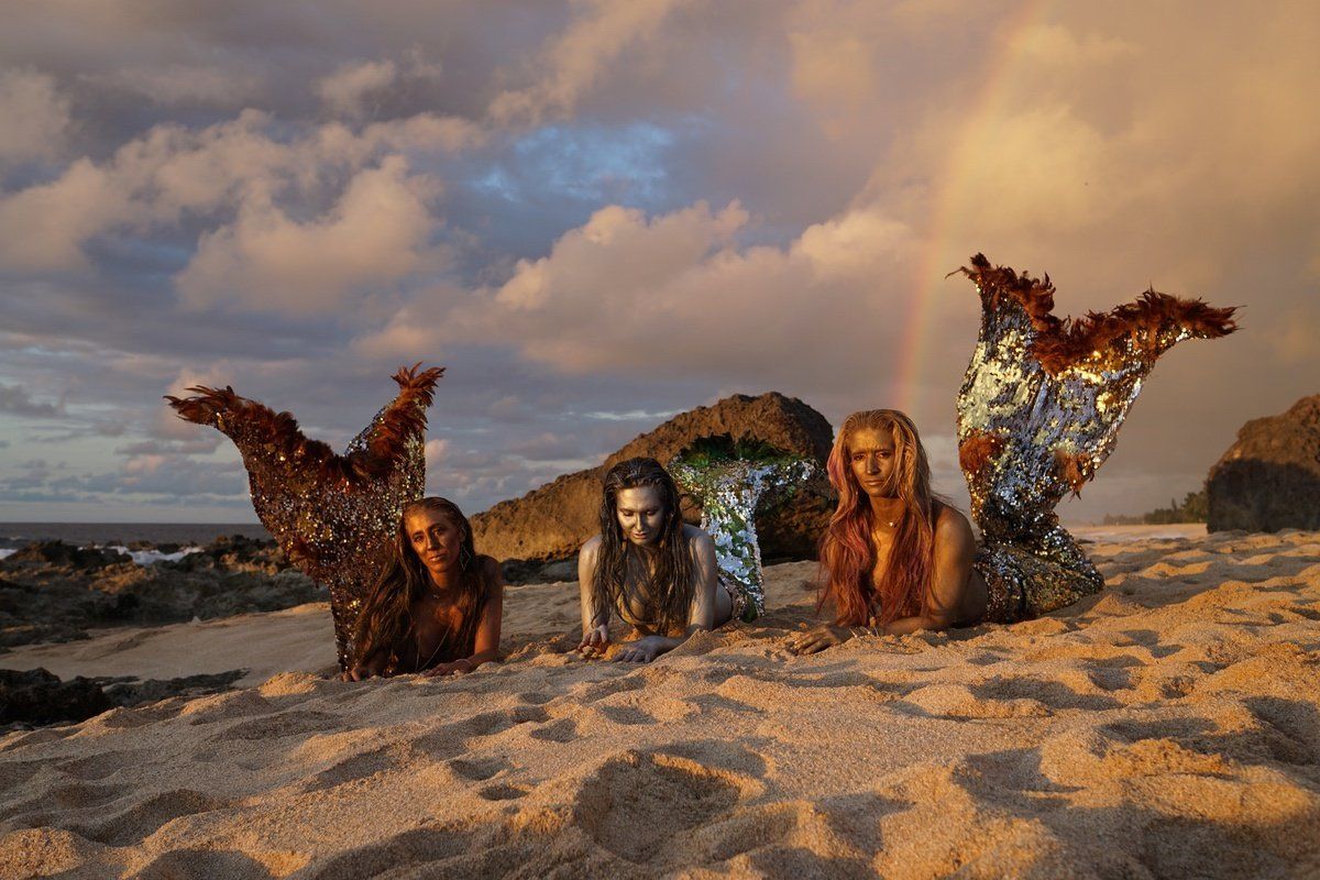 Three mermaids are laying on the beach with a rainbow in the background.