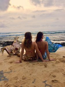 Two mermaids are sitting on the beach looking at the ocean.