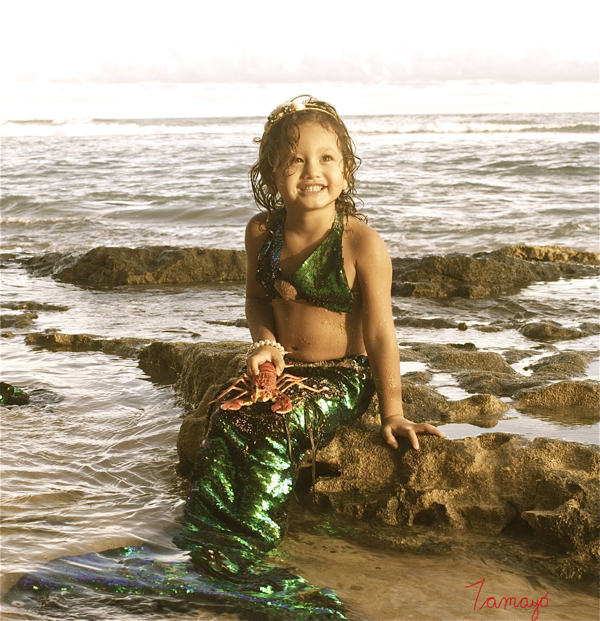 A little girl dressed as a mermaid is sitting on a rock near the ocean