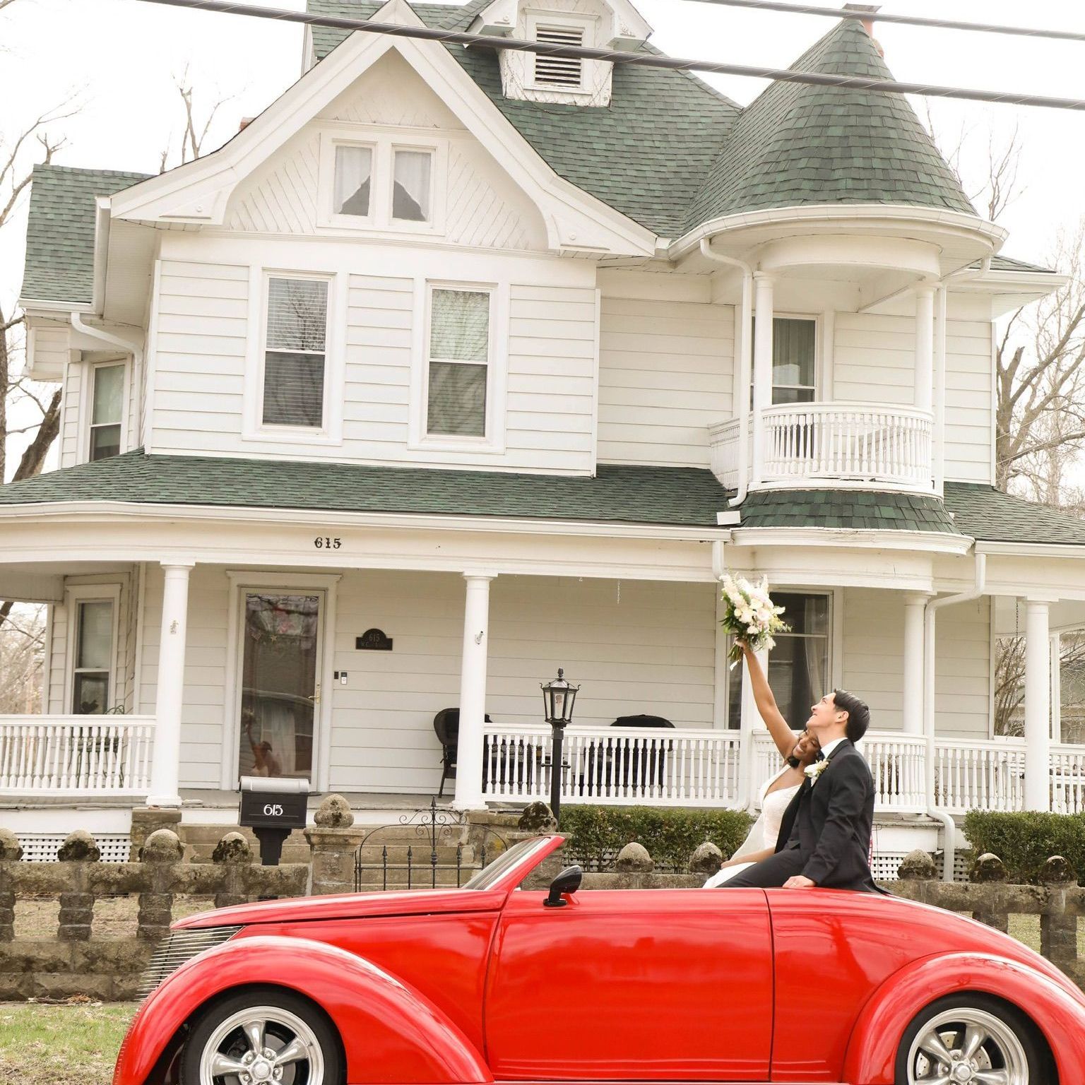 a bride and groom in a red car in front of a white house, smiling and waving
