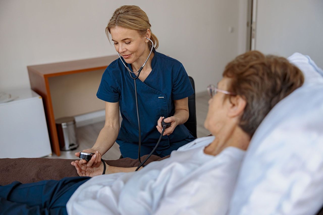 A healthcare professional in blue scrubs takes an older patient's blood pressure with a stethoscope and cuff in a medical setting.