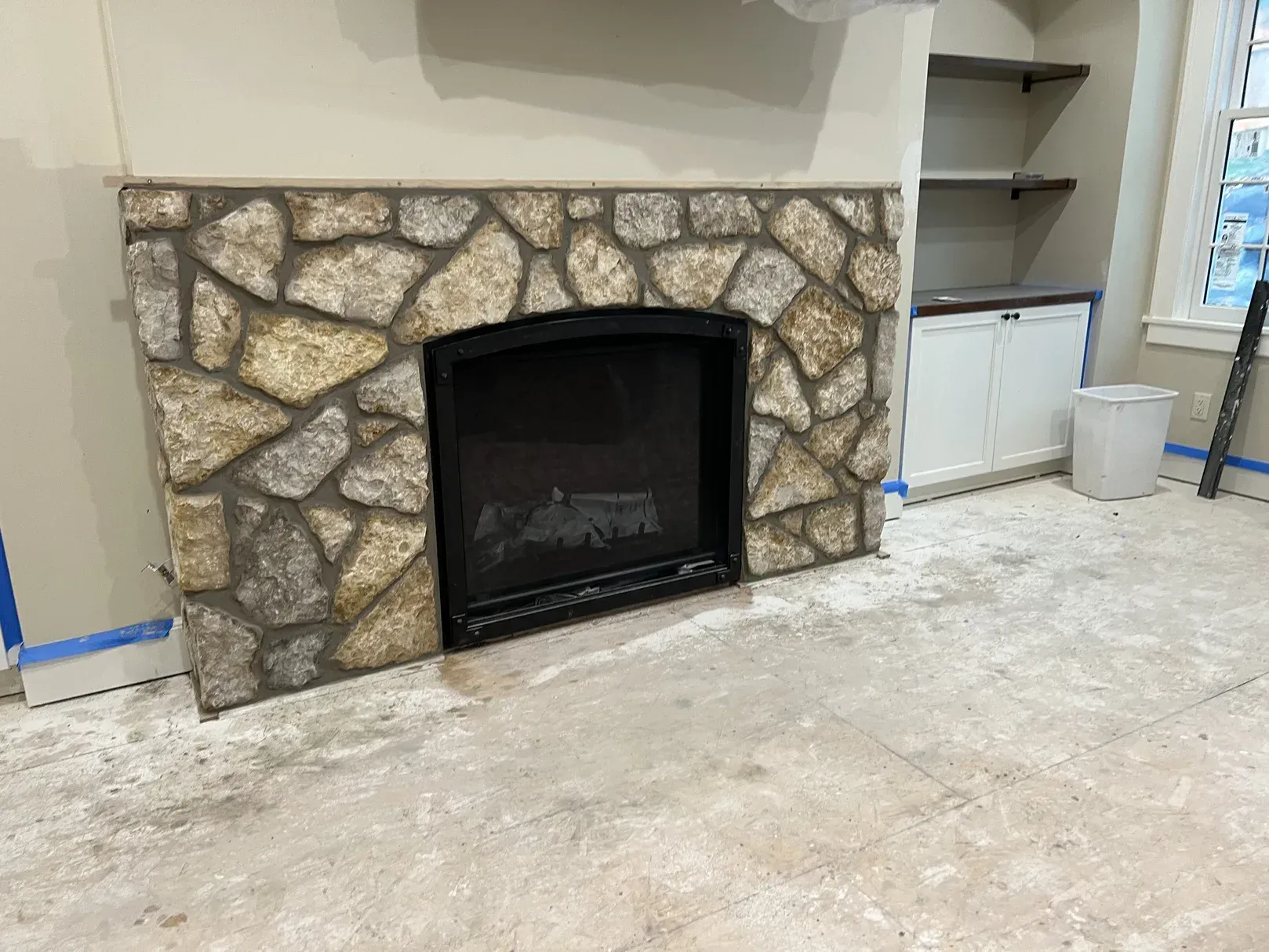 A stone fireplace in a living room with a ladder in the background.