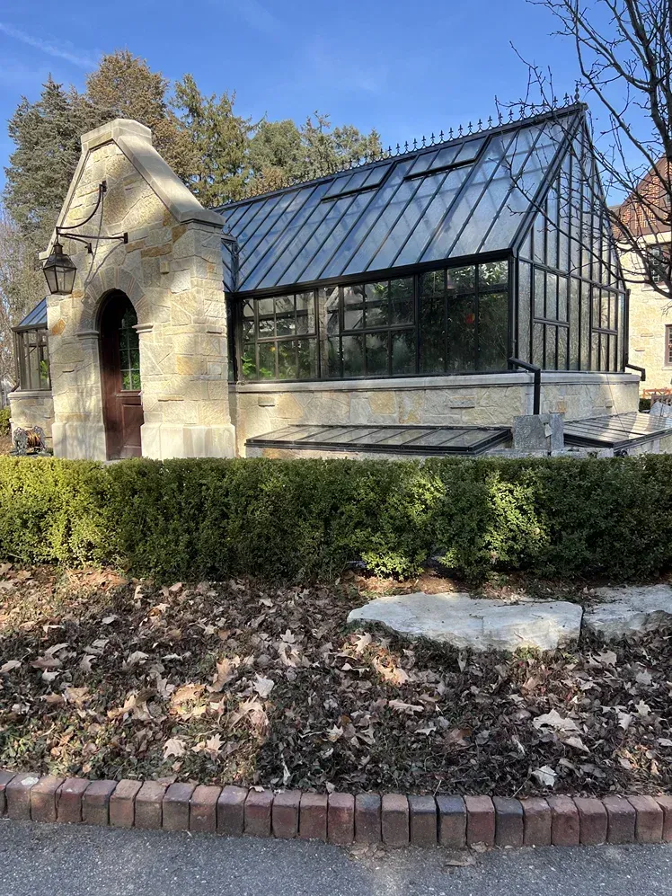 A greenhouse is sitting in the middle of a garden next to a stone building.