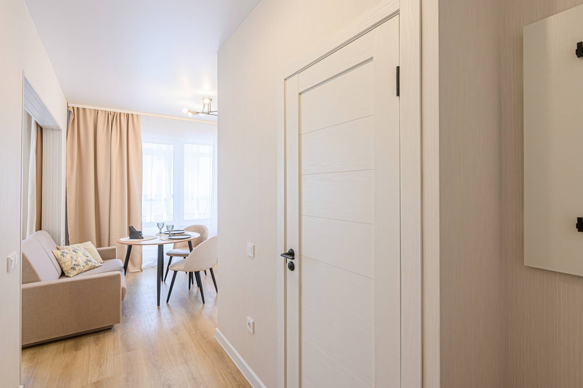 Beige hallway with a closed white door, leading to a living area with a sofa, table, and window with curtains.
