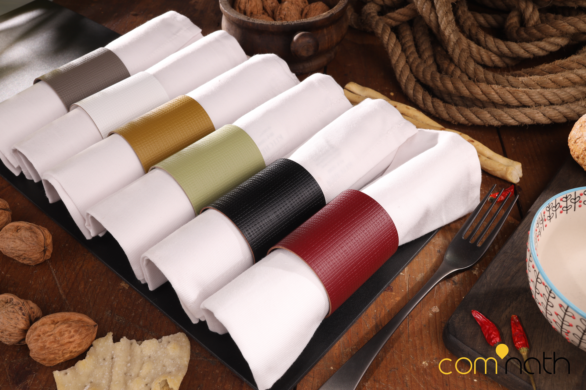 Serviettes blanches roulées avec des bandes de cuir colorées, disposées sur une table en bois avec de la nourriture et des couverts.