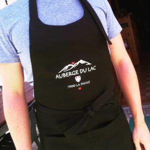 Personne portant un tablier noir avec le logo « Auberge du Lac » sur une chemise grise.