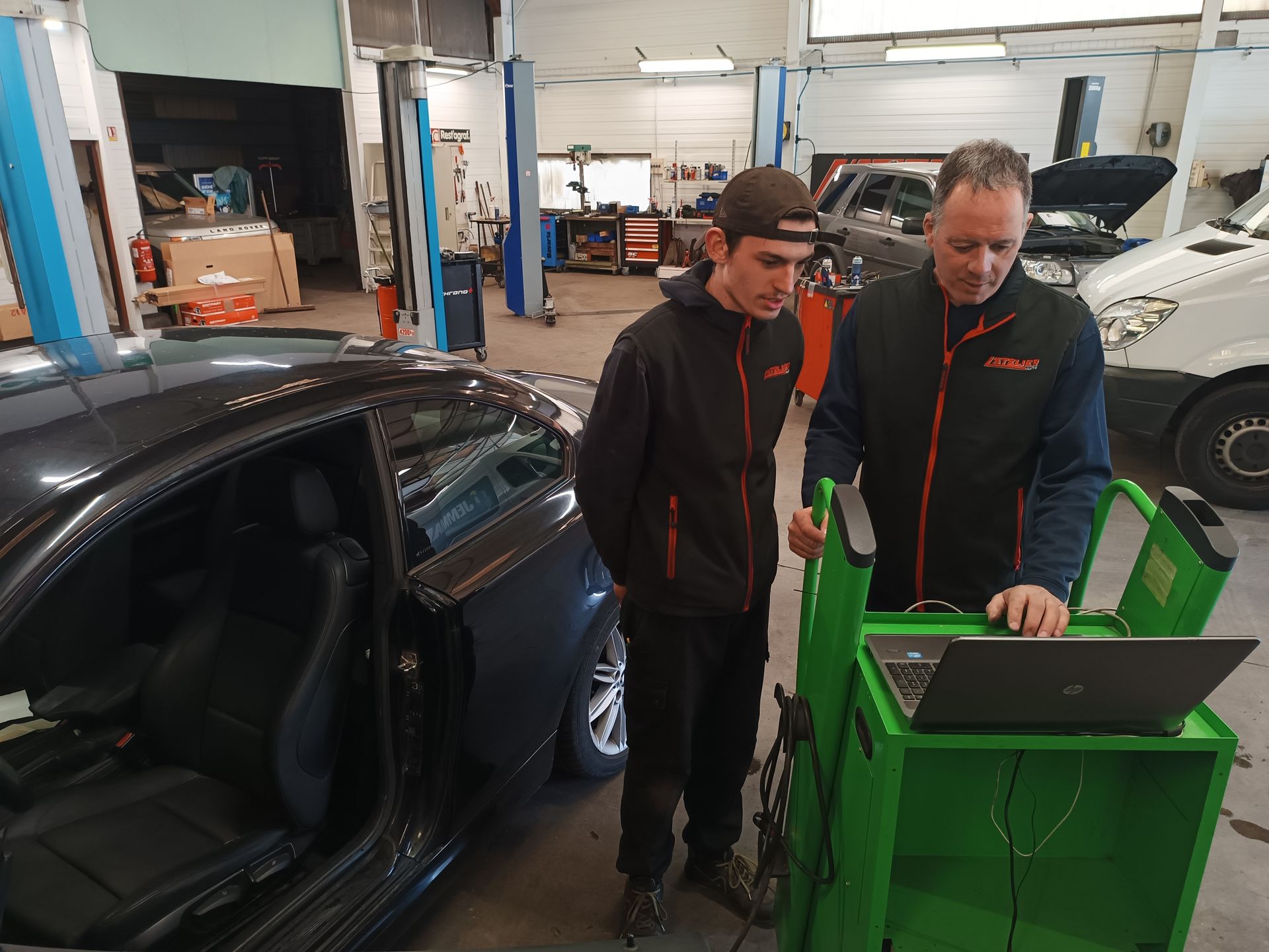 Deux hommes dans un garage regardent un ordinateur portable connecté à une voiture, probablement en train de diagnostiquer un véhicule.