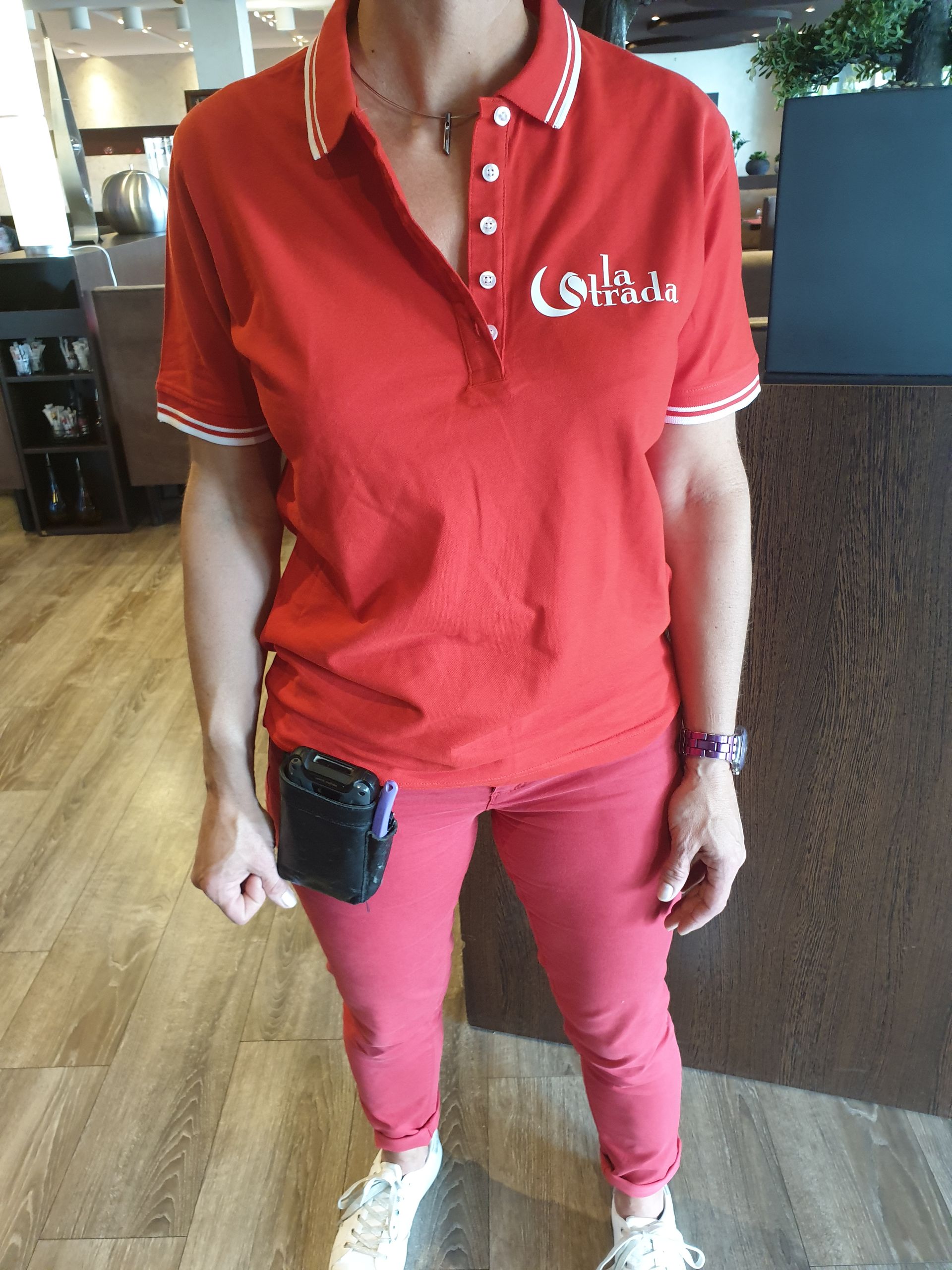 Personne en polo rouge et pantalon, sac banane noir, debout à l'intérieur près d'un comptoir.