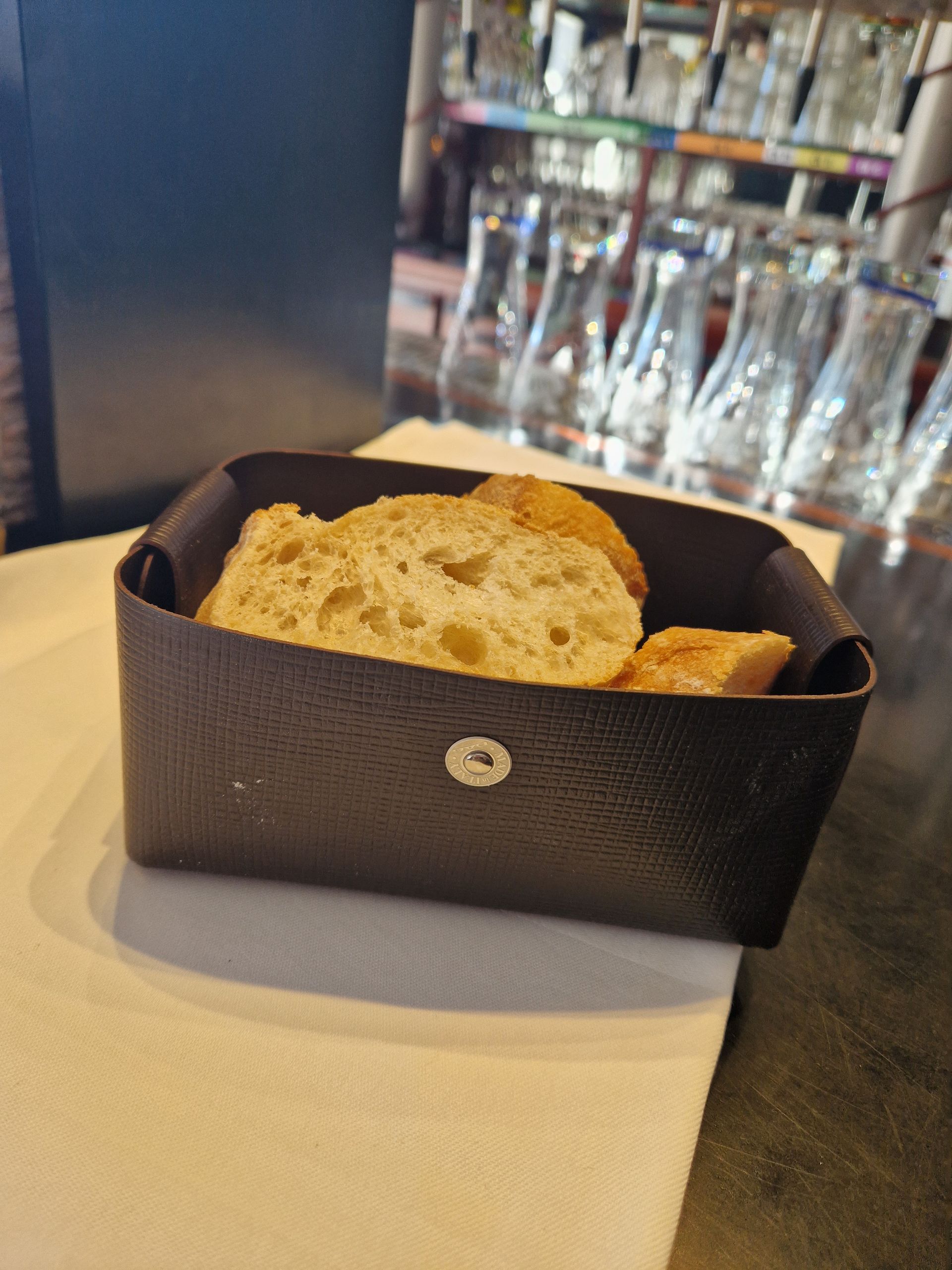 Du pain dans un panier marron foncé sur une serviette blanche, avec des verres et un bar en arrière-plan.