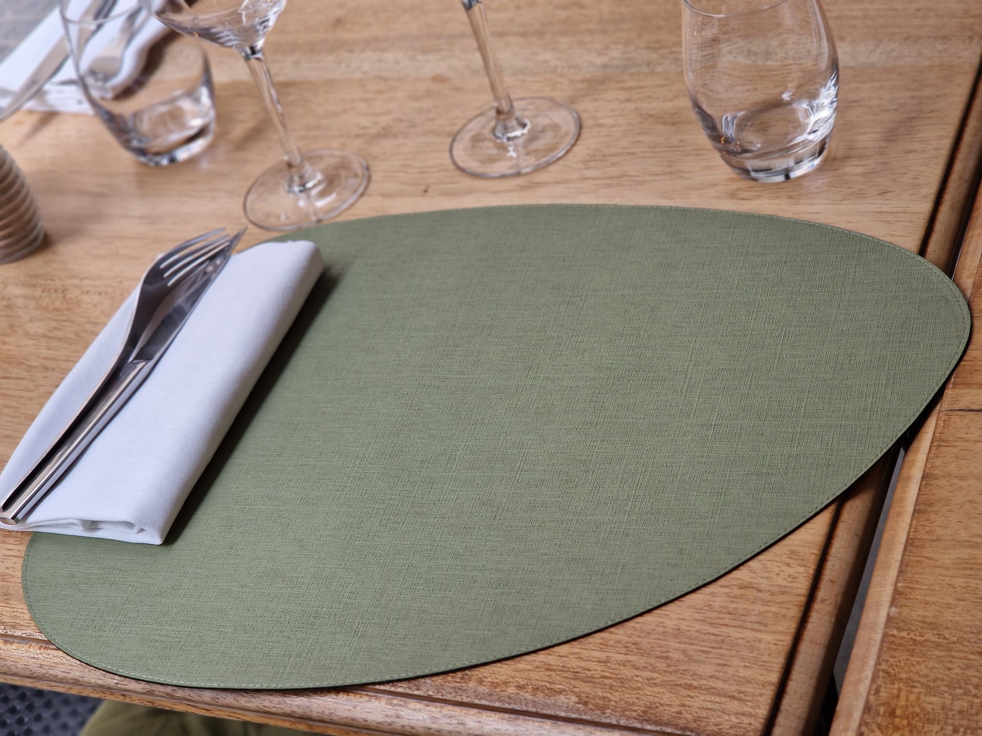 Set de table ovale vert sur une table en bois, avec couverts et serviette pliée.