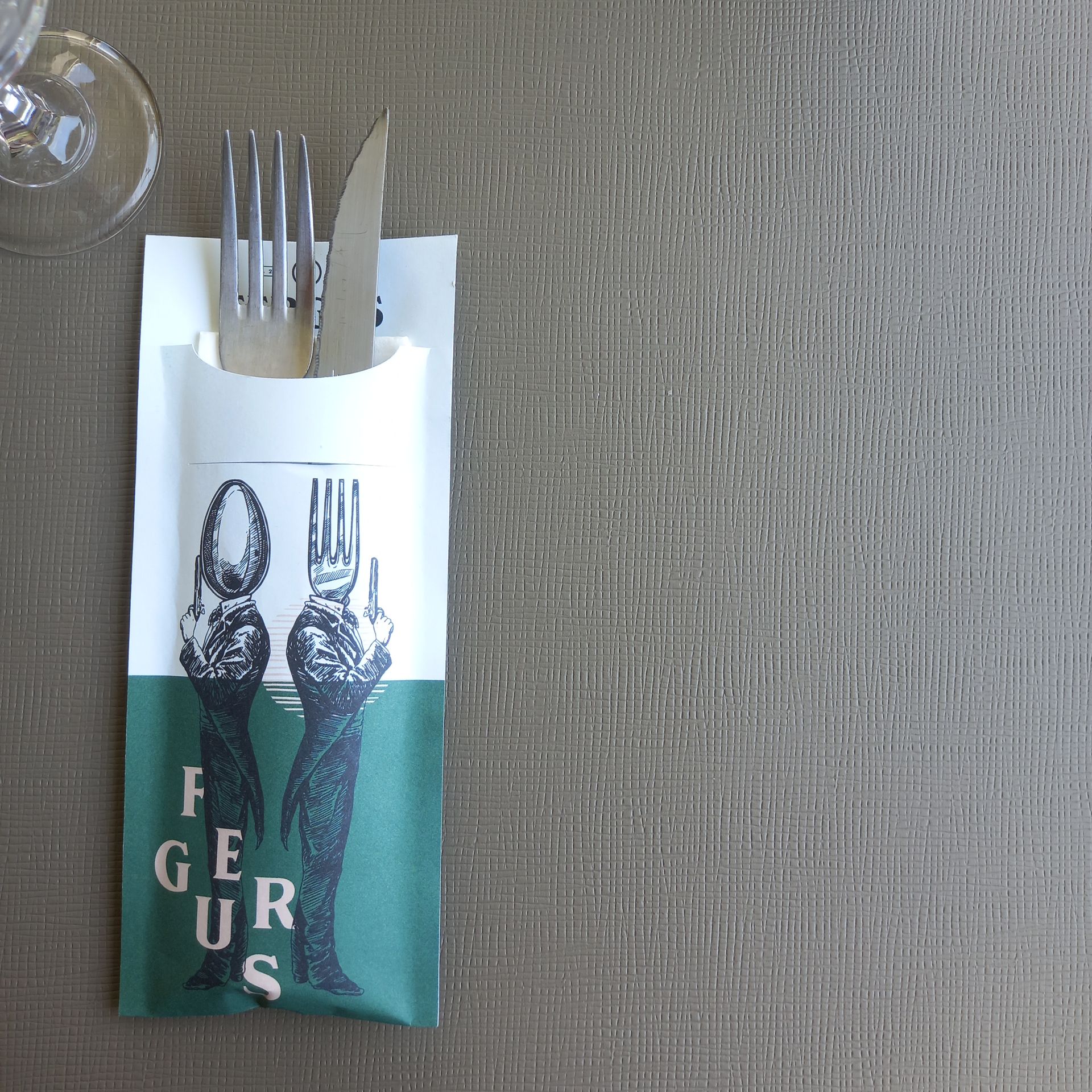 Fourchette et couteau dans une pochette en papier vert et blanc sur un set de table gris texturé. Un verre à vin est placé en haut à gauche.