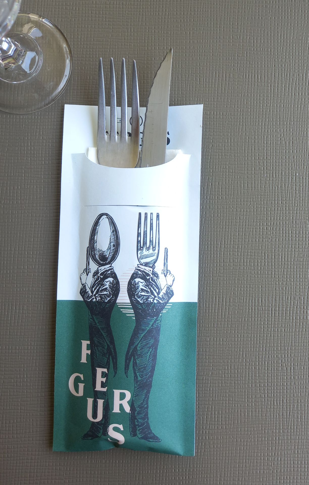 Un porte-ustensiles en papier avec une fourchette et un couteau à l'intérieur ; vert et blanc avec des figures fantaisistes.