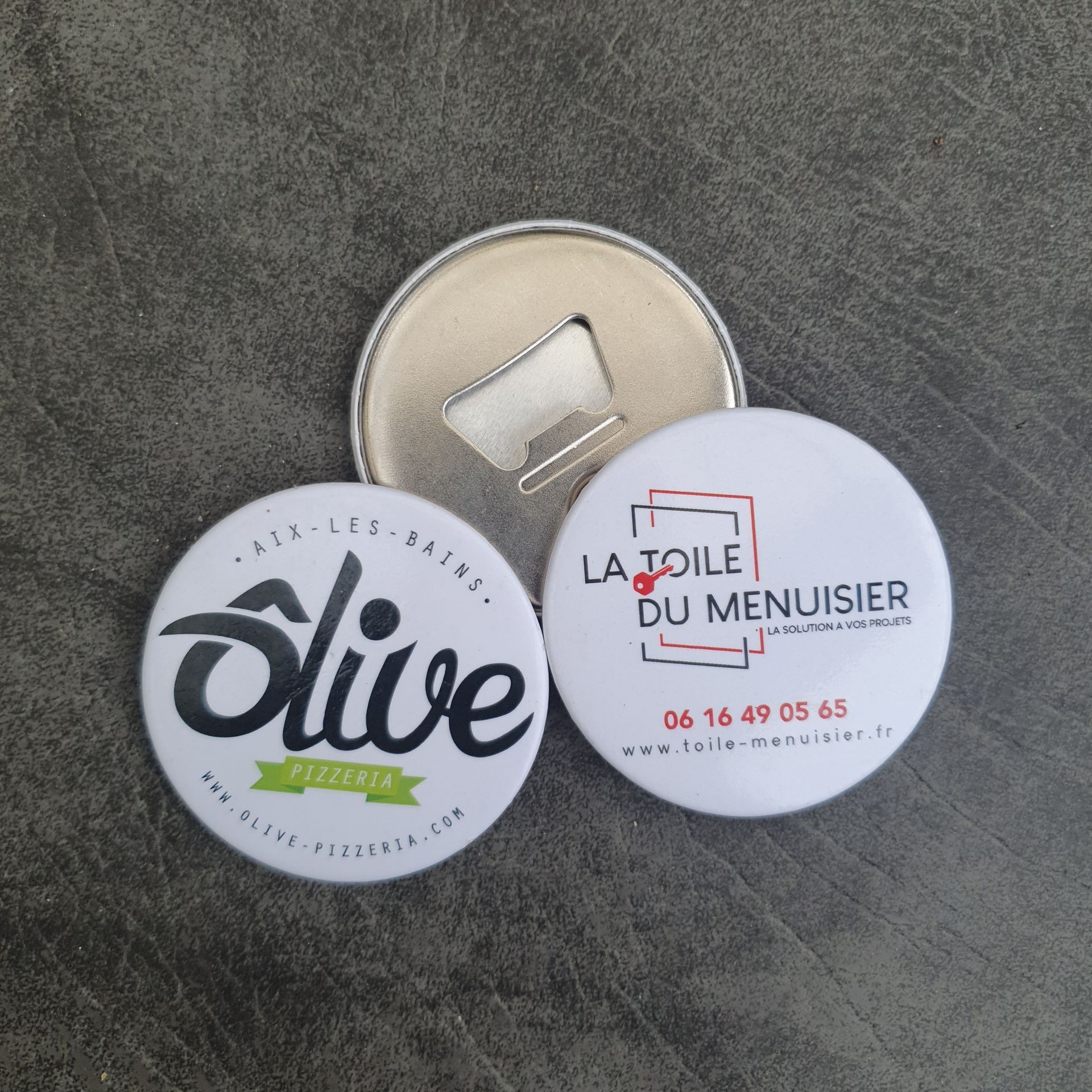 Décapsuleur avec deux boutons ronds blancs comportant des logos d'entreprise sur une surface grise.