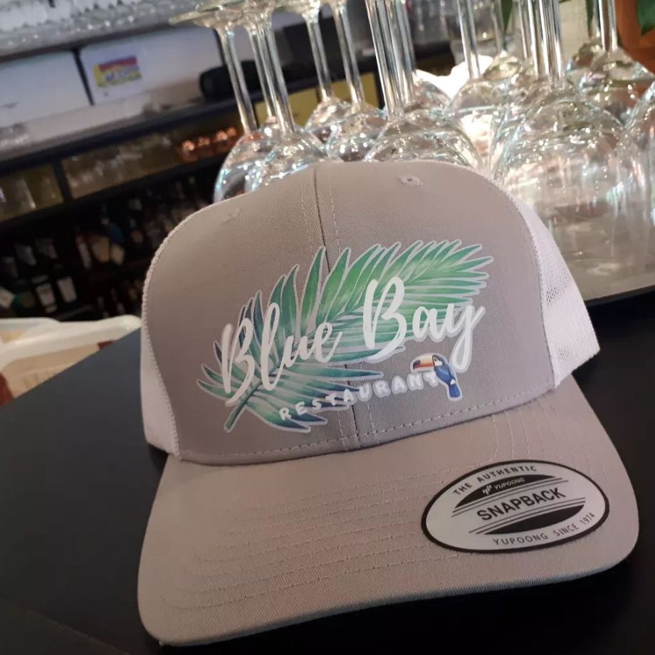 Casquette de baseball grise et blanche avec le logo « Blue Bay Restaurant », posée sur une table dans un restaurant.
