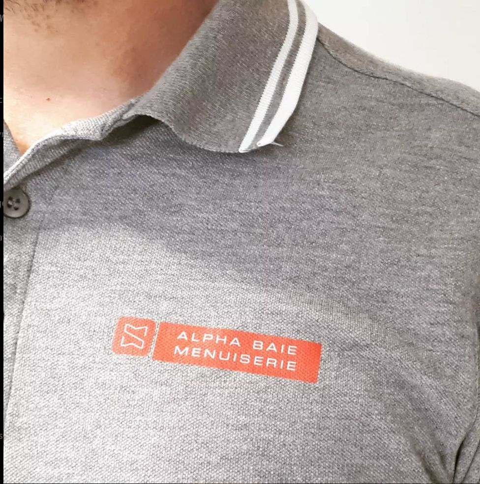 Homme portant un polo gris à rayures blanches sur le col. Logo orange « Alpha Baie Menuiserie » sur la poitrine.