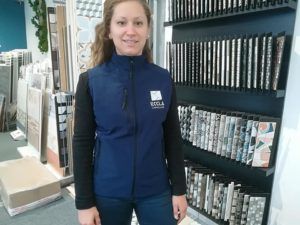 Une femme en gilet bleu se tient debout dans une salle d'exposition de carrelage, souriante. Des échantillons de carrelage sont disposés sur les étagères.