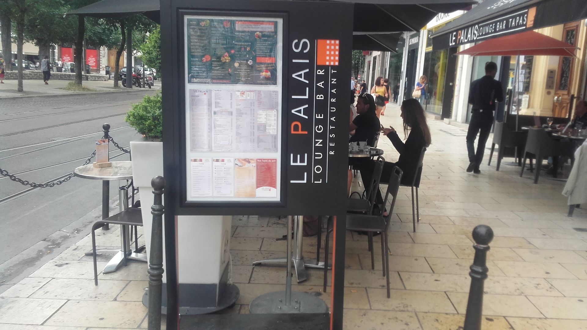 Restaurant en plein air : panneau « Le Palais Bistro & Bar », clients assis à des tables.
