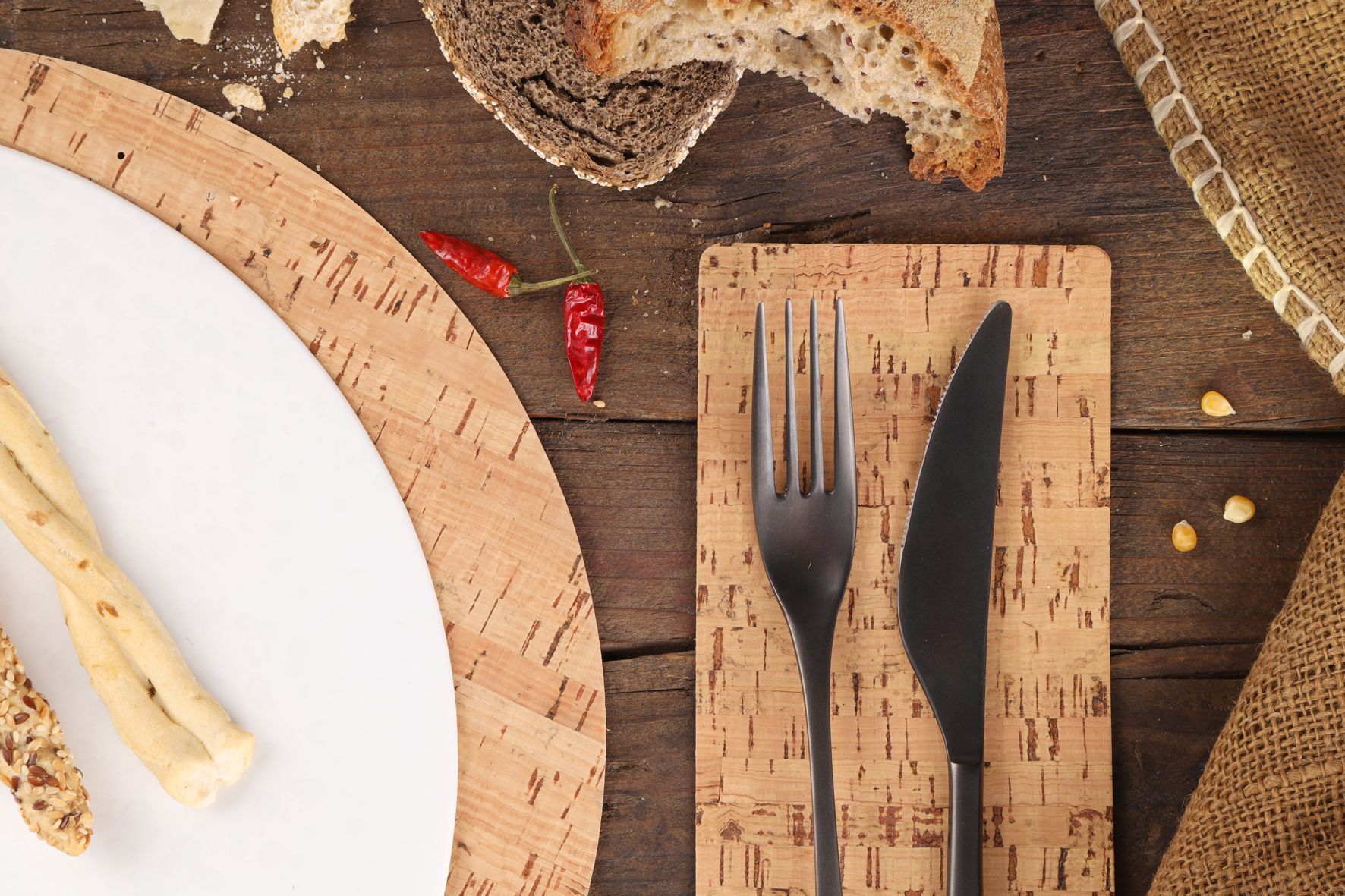 Décoration de table rustique : assiette blanche, gressins, fourchette, couteau sur set de table en liège, pain, piments rouges.