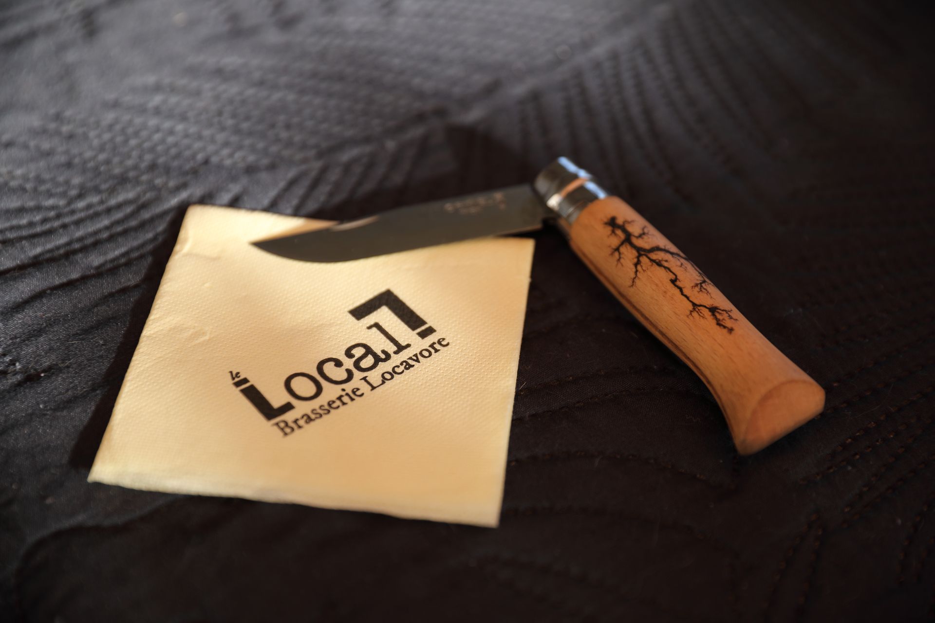 Un couteau de poche repose sur une serviette portant le logo « Local Brasserie Lockport ». Fond gris foncé.