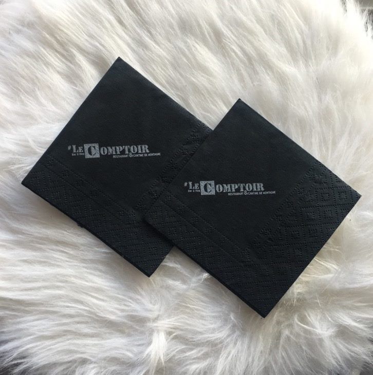 Deux serviettes à cocktail carrées noires avec logo blanc, sur une surface en fourrure blanche.