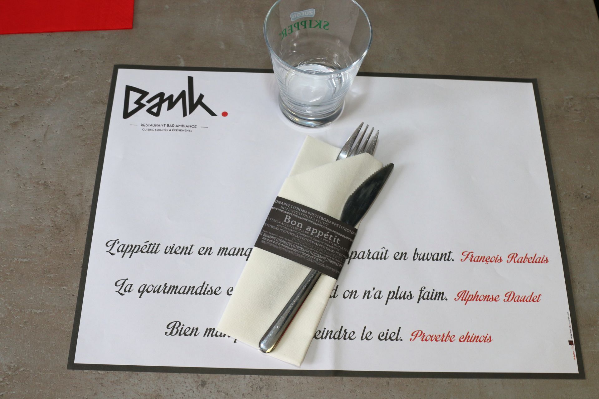 Couvert au restaurant « Bank », avec serviette, couverts, verre d'eau et menu sur un set de table.
