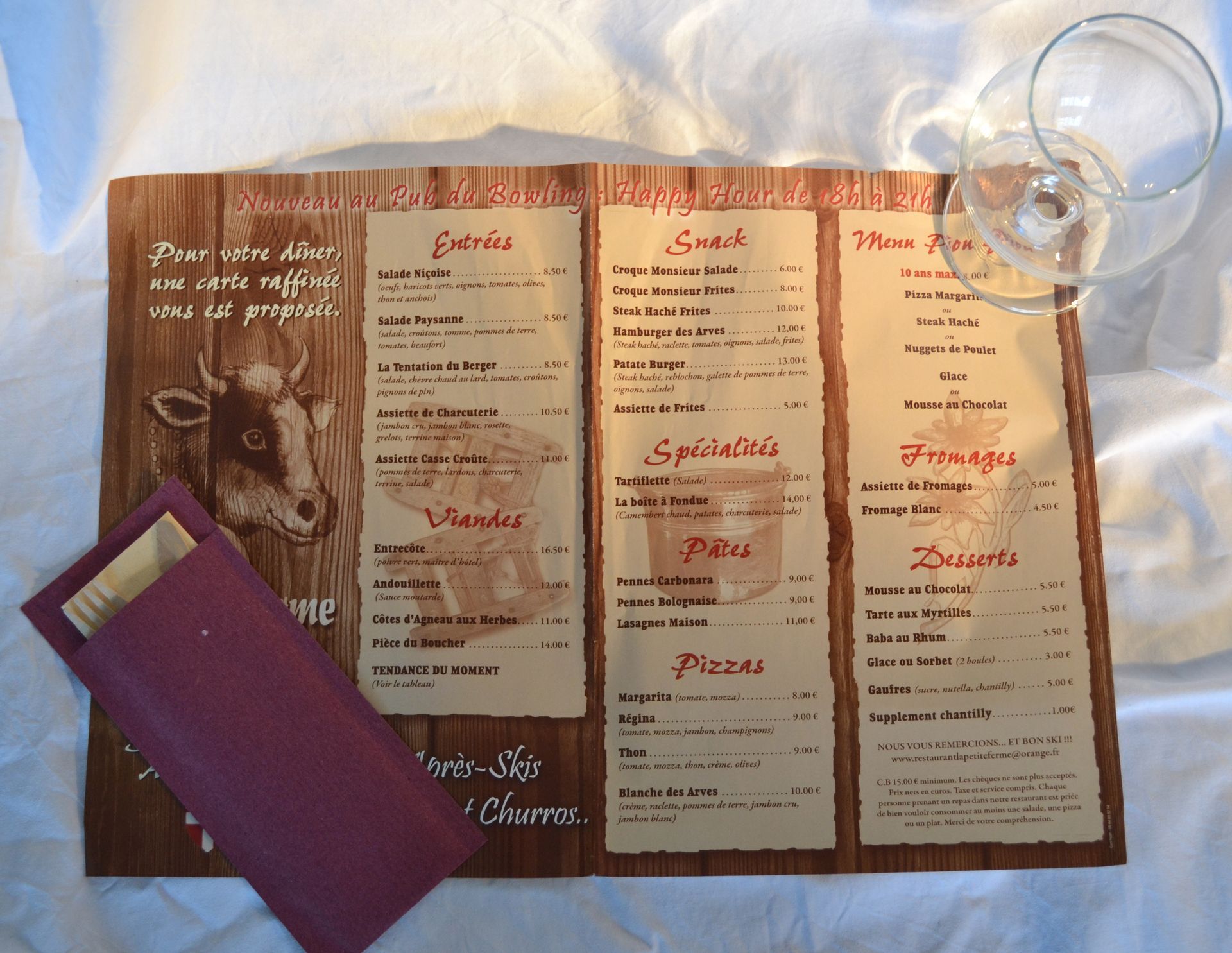 Menu affiché sur une surface en bois avec une illustration de vache, un porte-serviettes violet et deux verres à vin vides.