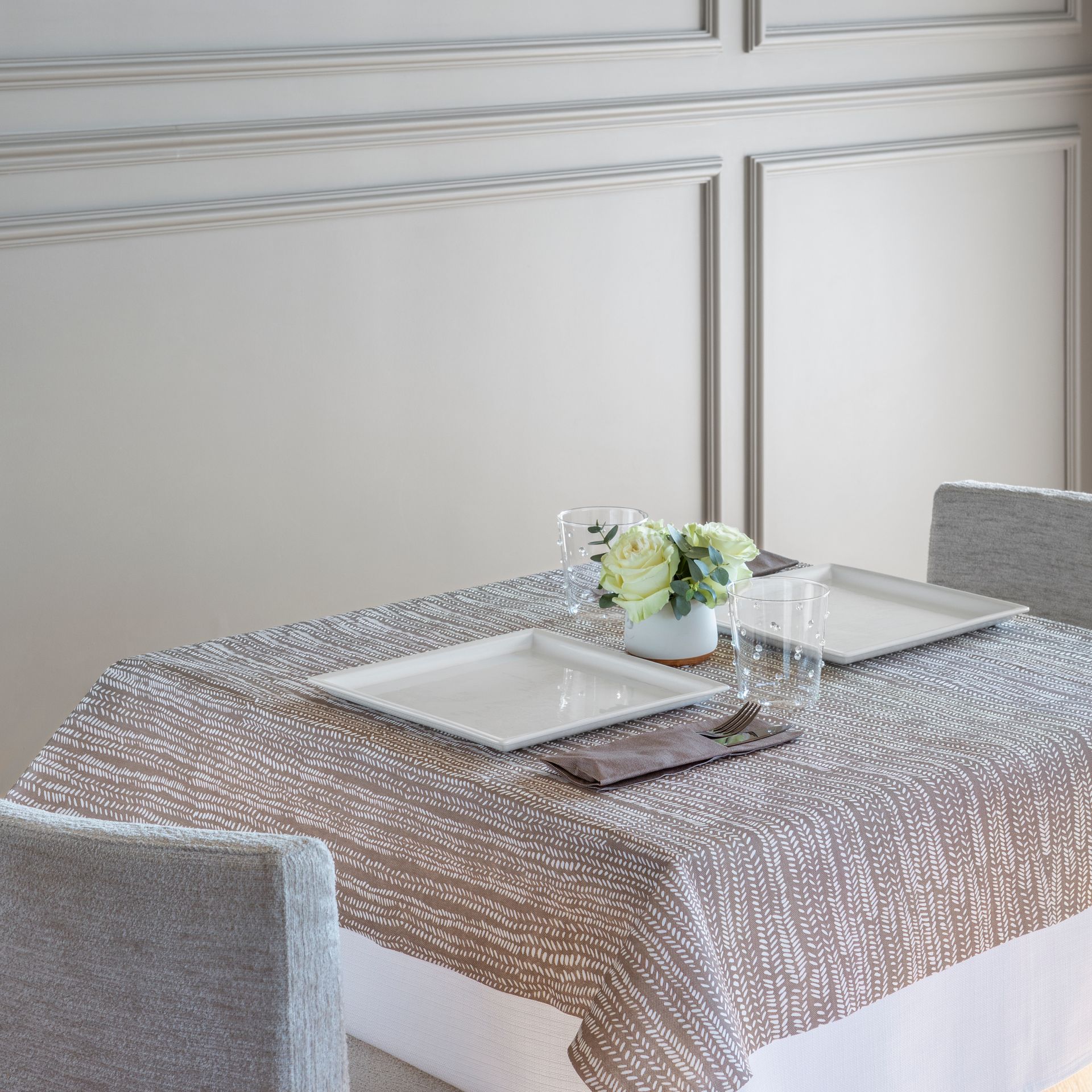 Une table dressée pour deux personnes avec des assiettes blanches et un centre de table fleuri. On aperçoit des chaises grises et une nappe à motifs.