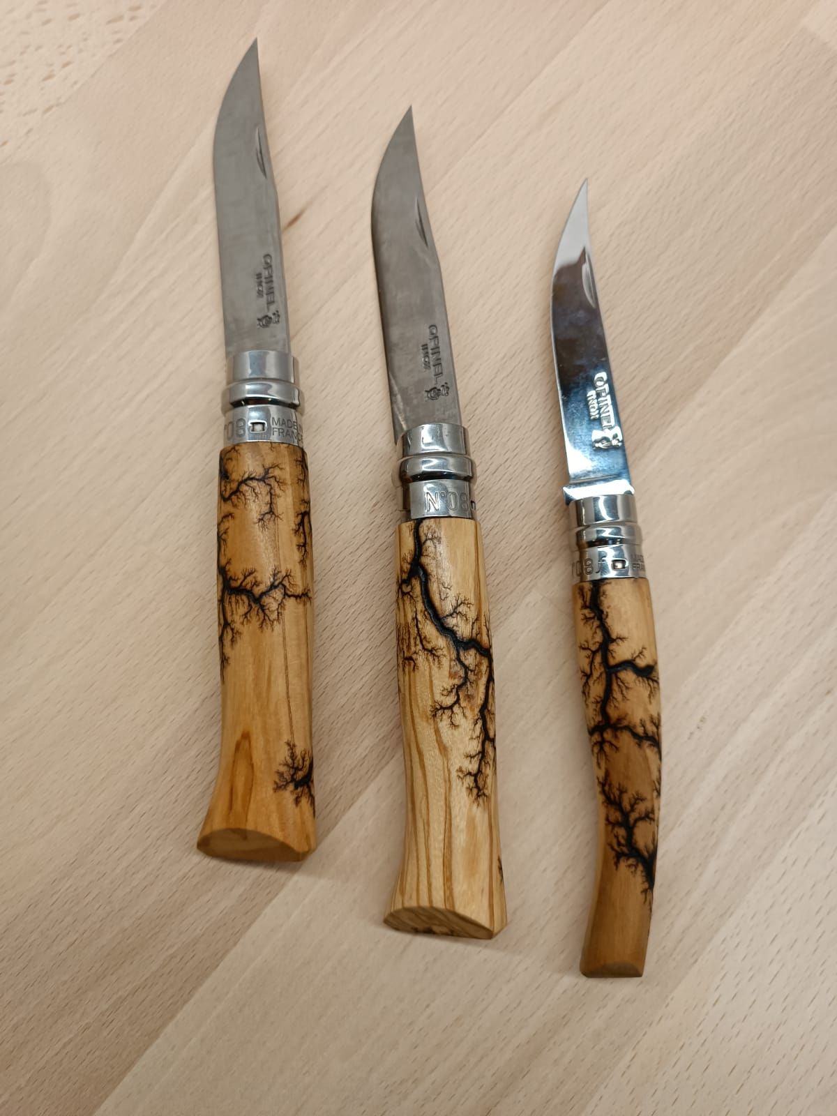 Trois couteaux de poche avec manches en bois, chacun gravé d'un motif d'éclair, reposant sur une surface en bois clair.