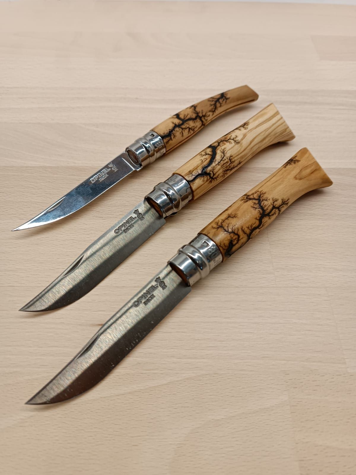 Trois couteaux de poche avec manches en bois sur une surface en bois clair. Chaque lame est partiellement ouverte.