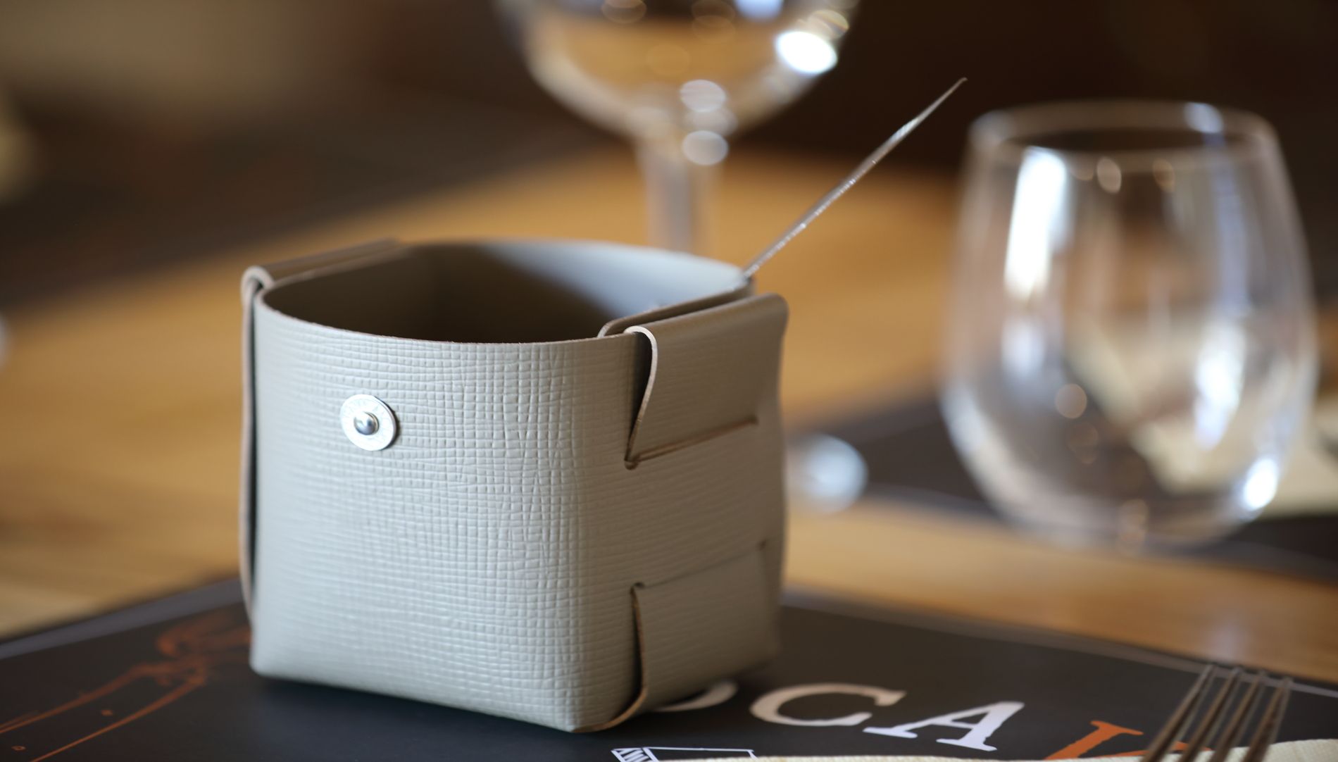 Un petit récipient en cuir beige avec une aiguille posé sur une table près de verres à vin vides.