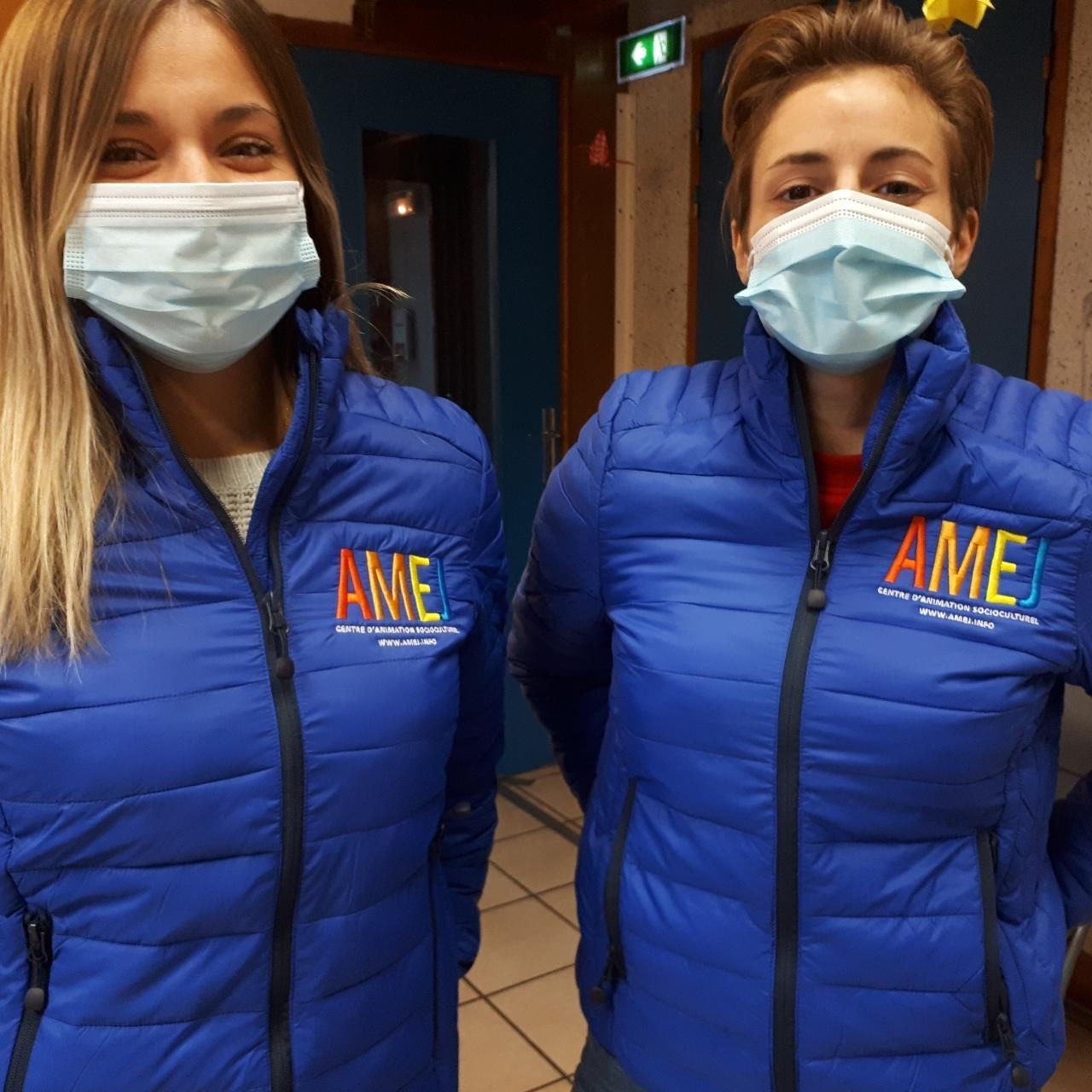 Deux femmes portant des vestes bleues et des masques, debout à l'intérieur. Les deux vestes arborent le logo « AMEJ ».