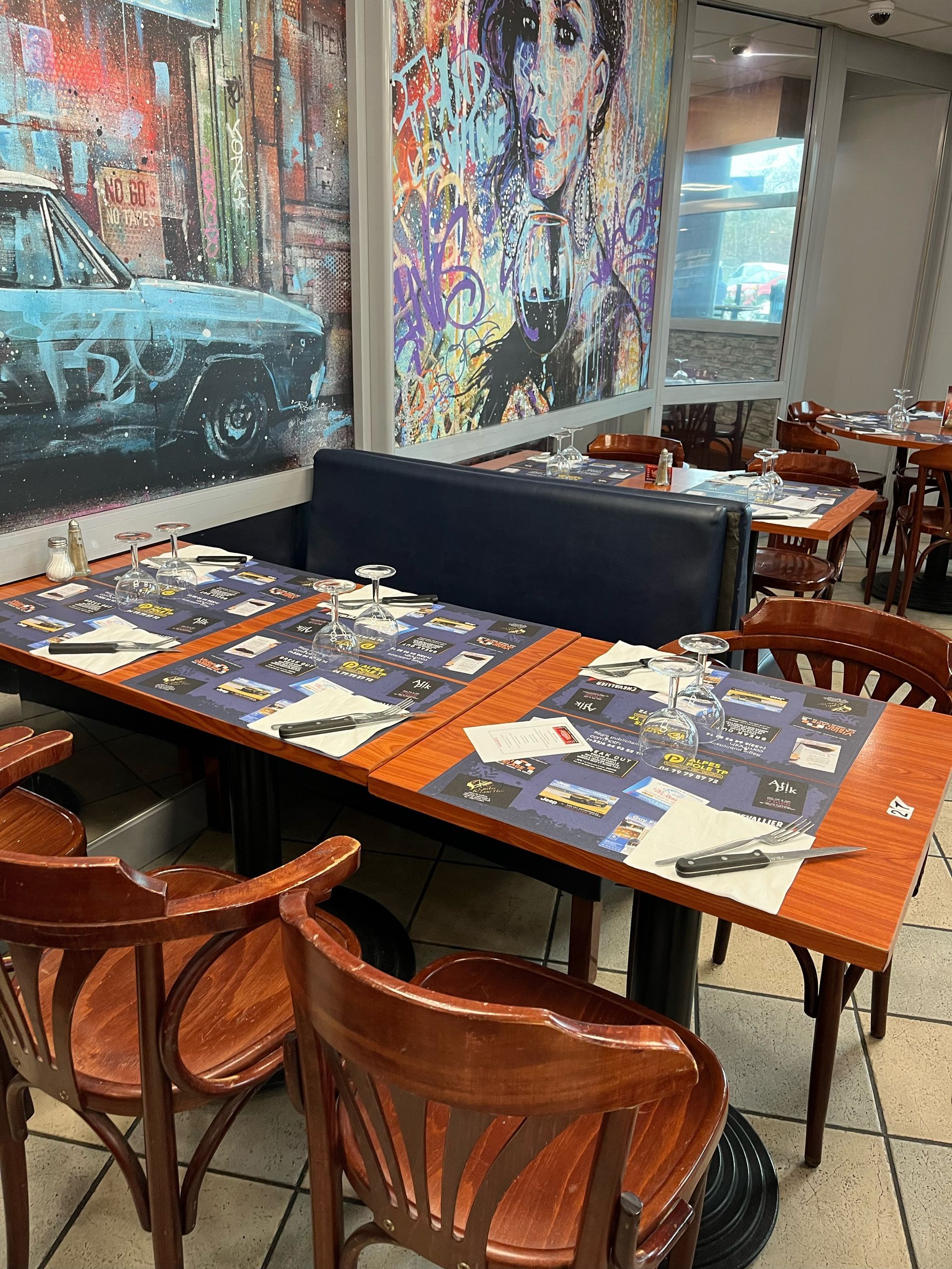 Espace salle à manger du restaurant avec tables dressées, sets de table orange, chaises en bois, peintures murales colorées sur les murs.