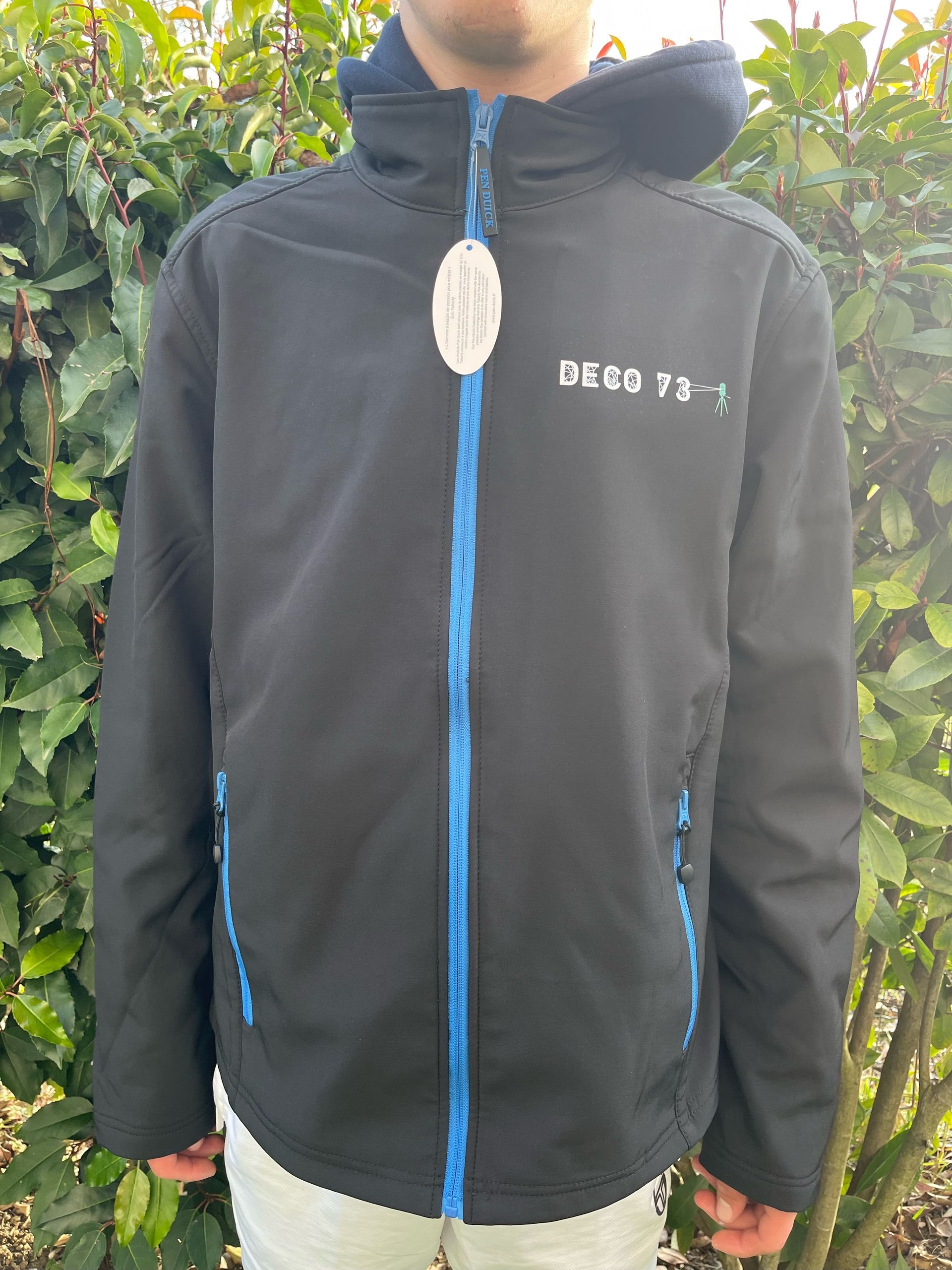 Garçon portant une veste noire avec fermeture éclair bleue, étiquette blanche et capuche, debout à l'extérieur.
