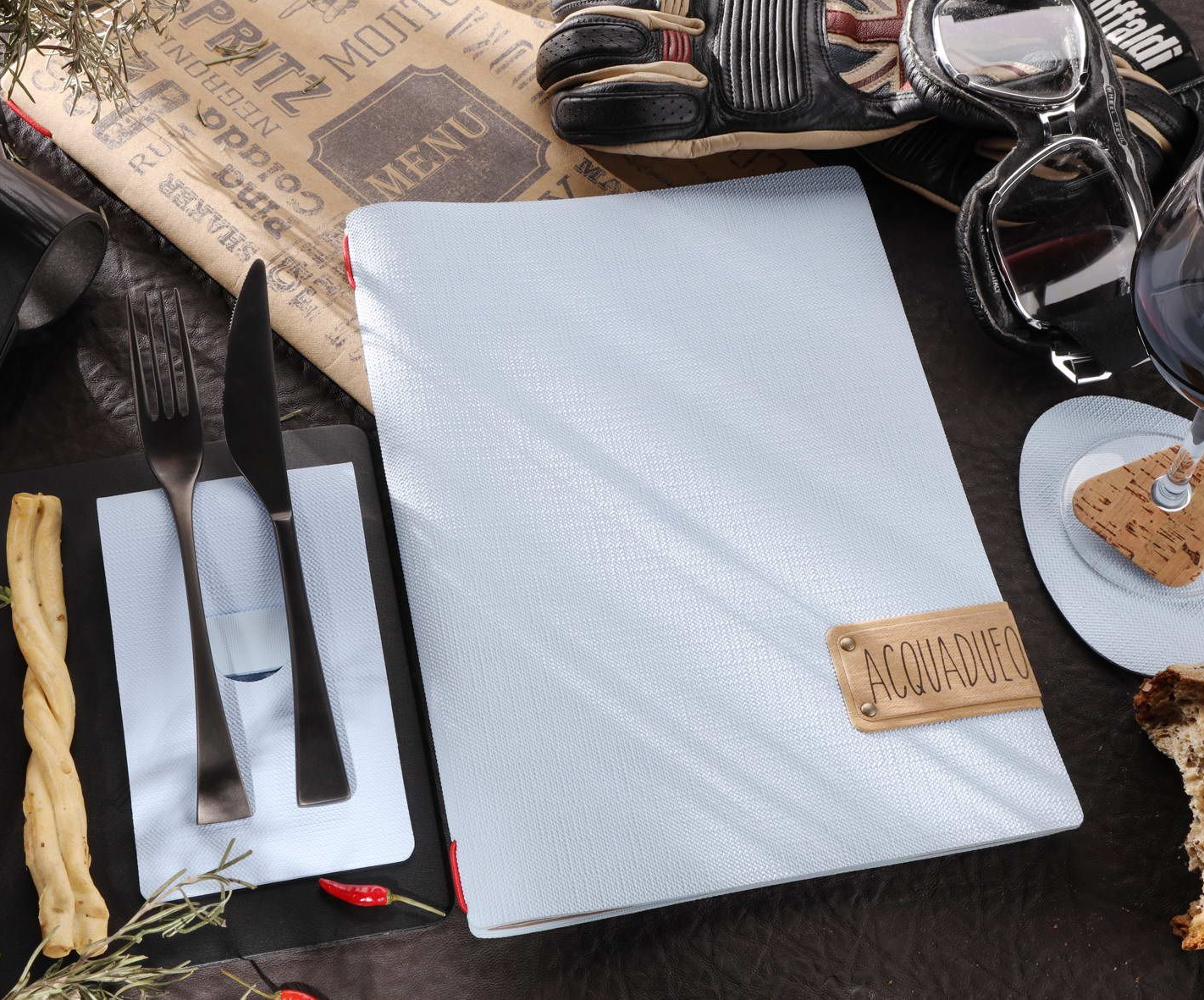 Cadre de restaurant avec un menu, des couverts, des gressins et un verre de vin sur une surface sombre.