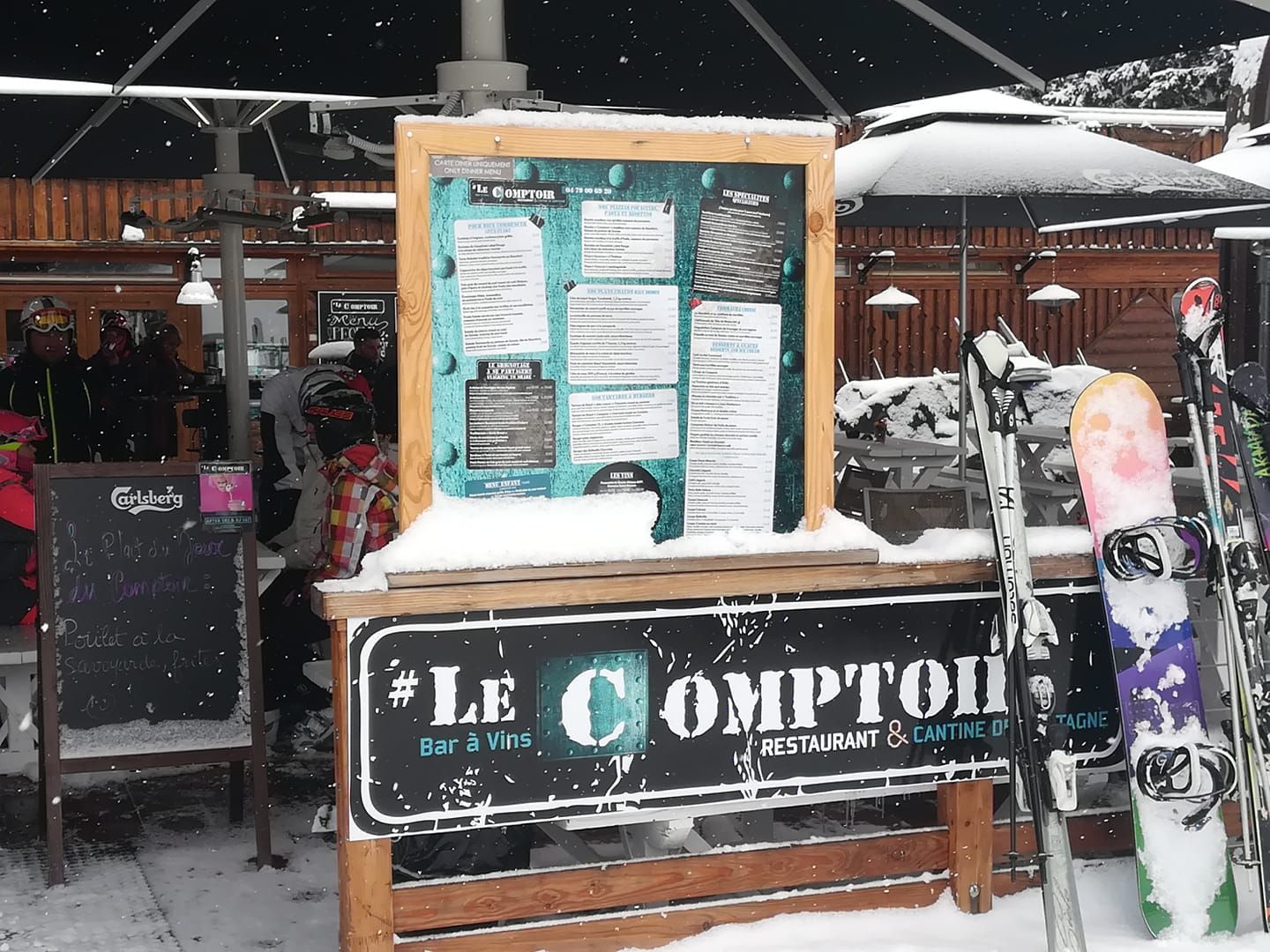 Café extérieur enneigé « Le Comptoir » avec tableau de menu, skis et clients.